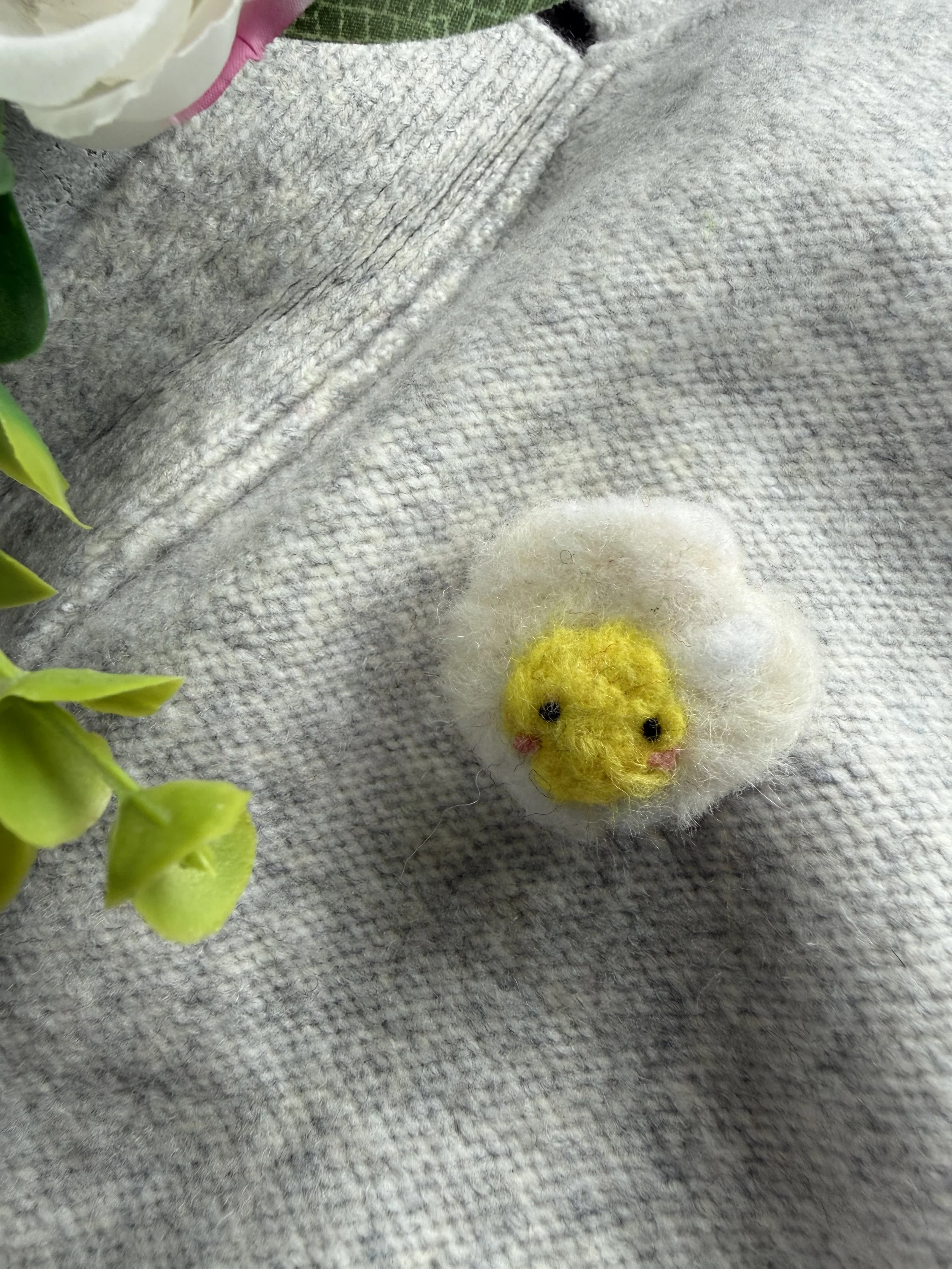 Close-up of a small, felted egg pin, on a gray fabric surface, with some green leaves in the foreground and a nearby white and pink flower.