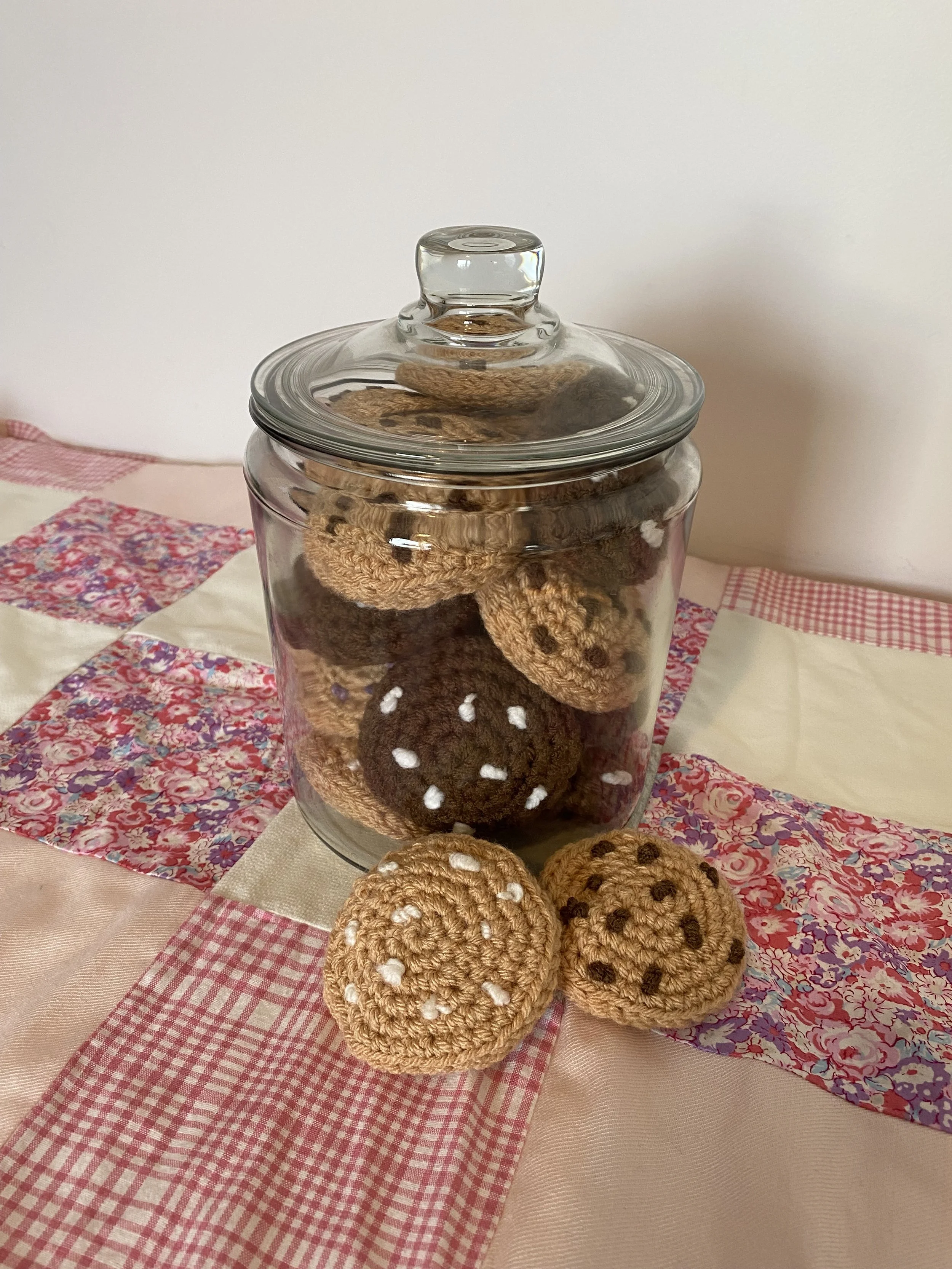 A glass jar with a lid containing crochet cookies with white and dark brown yarn in a patchwork cloth setting.
