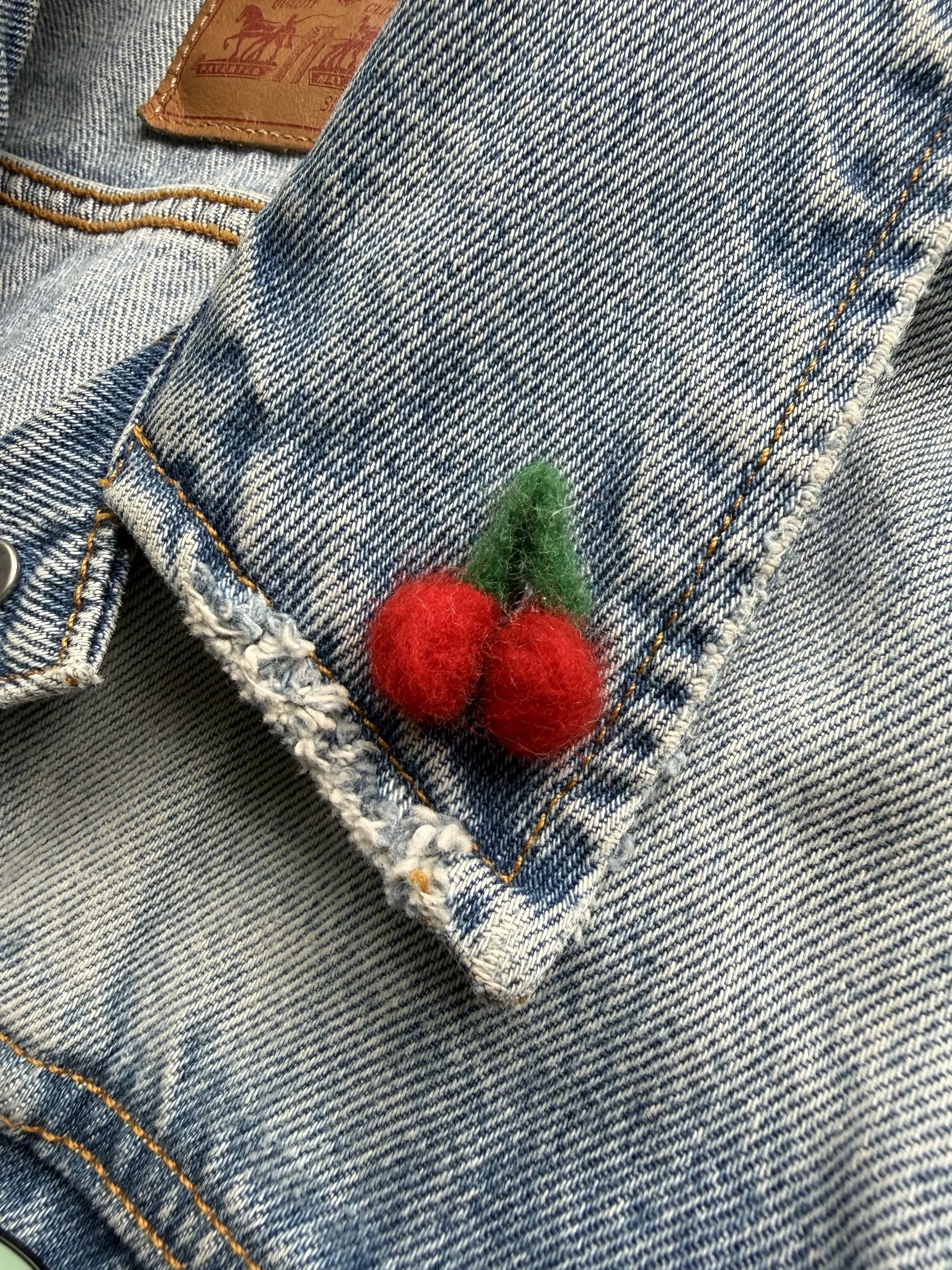 Close-up of denim jeans with a small red and green felted cherry pin.