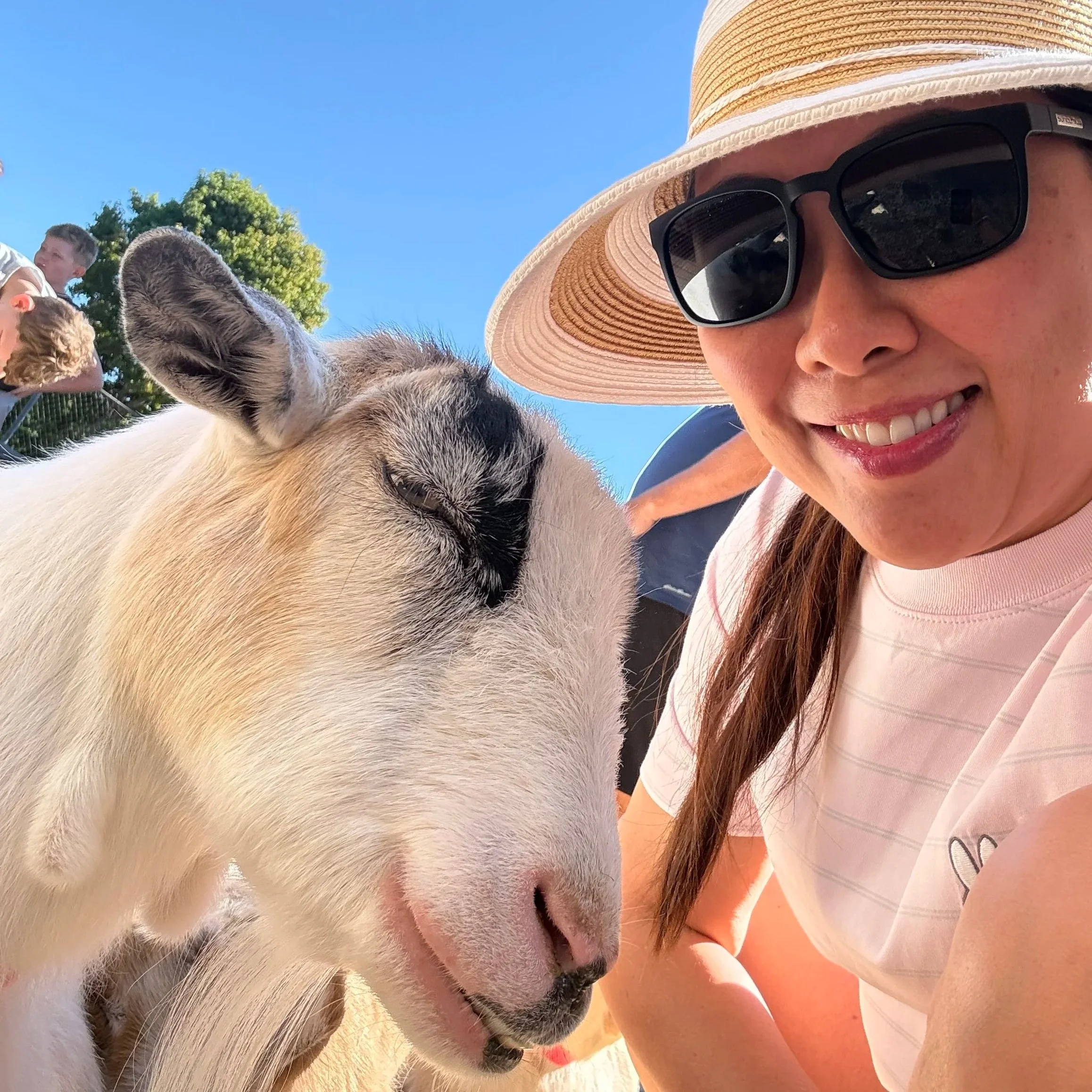 A woman smiling and wearing a wide-brimmed straw hat and sunglasses, close to a goat with its eyes closed, outdoors on a sunny day.