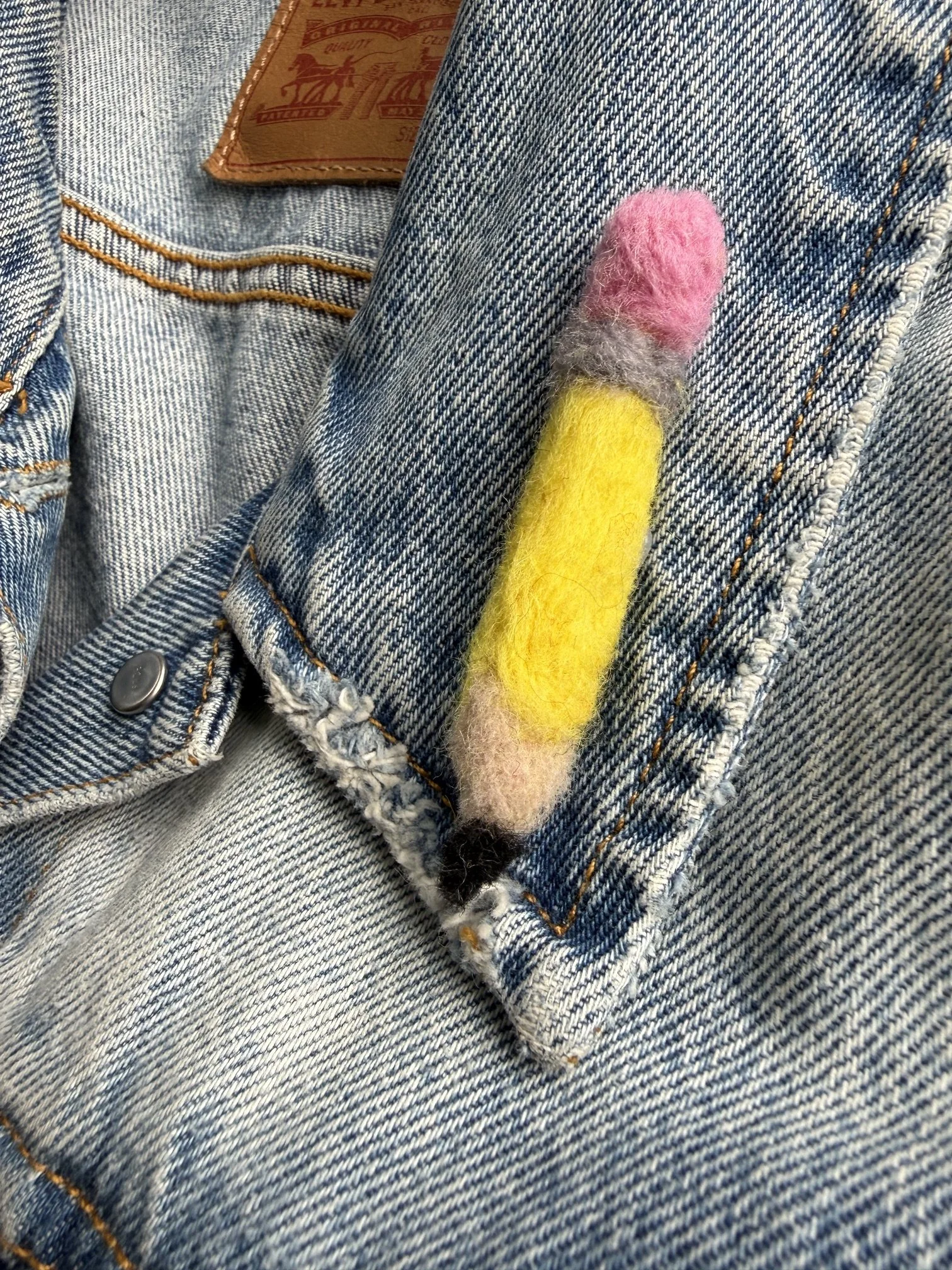 Close-up of a denim jacket with a felted pencil pin attached to the pocket.
