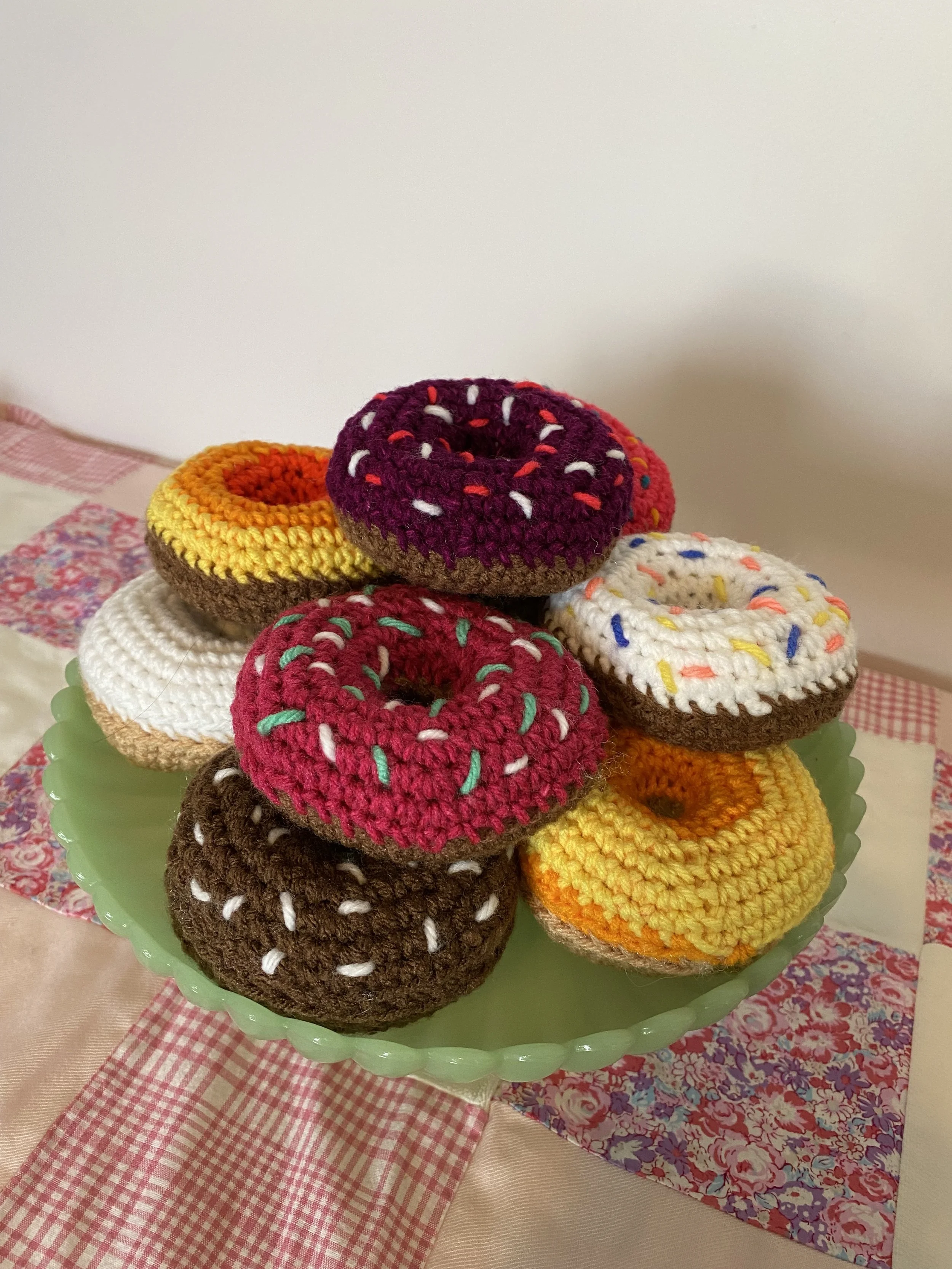 A green serving dish holding multiple colorful crocheted donuts with sprinkles, set on a table with pink and floral tablecloths.