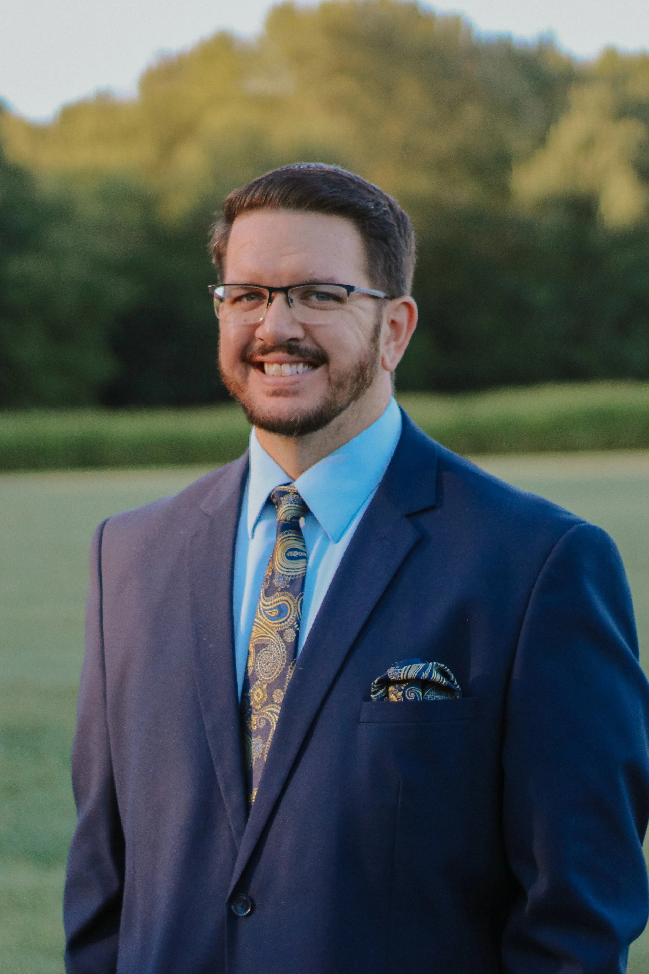 Smiling man wearing glasses, a navy suit, blue shirt, and paisley tie outdoors