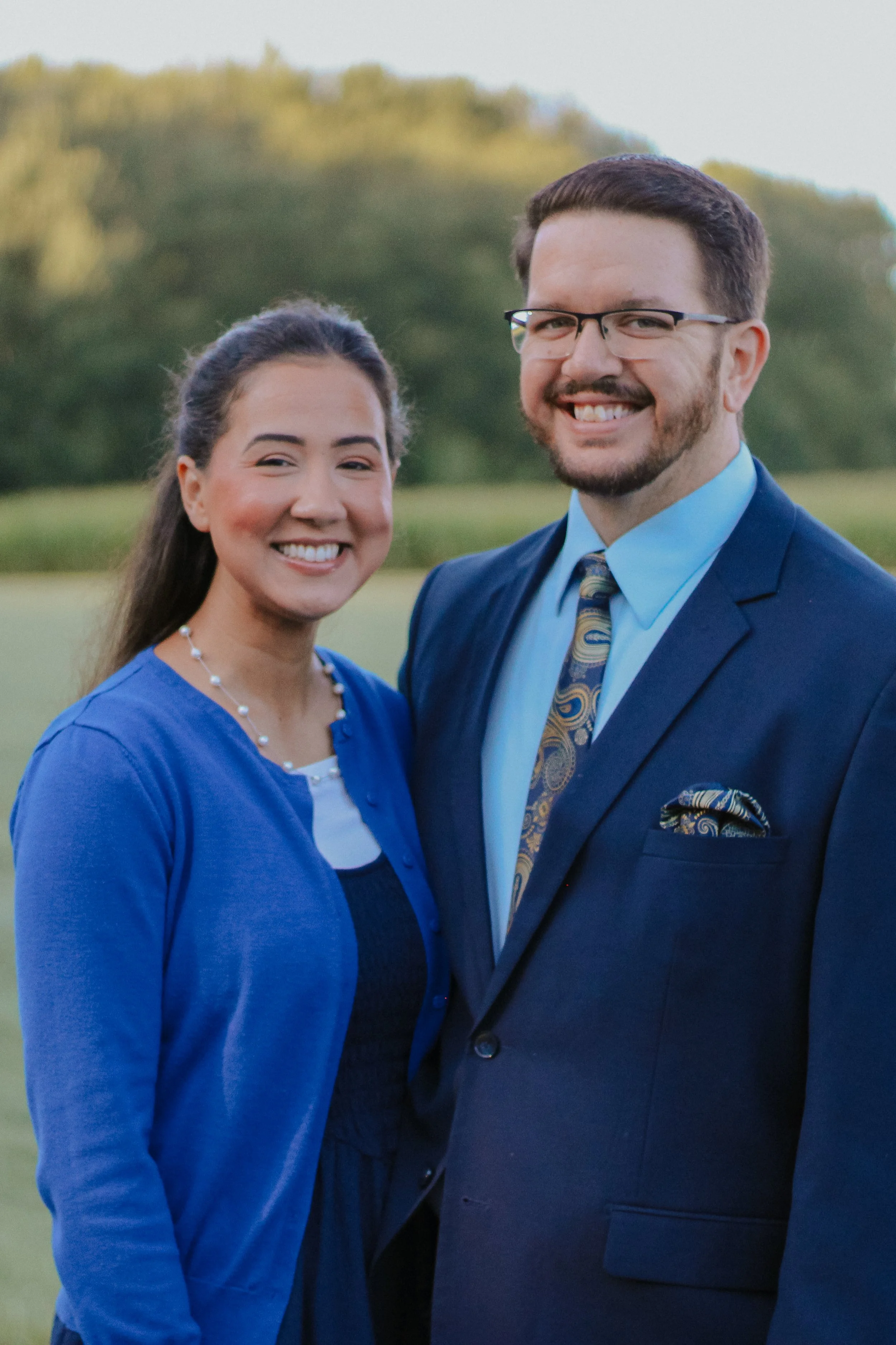 Smiling couple in formal attire posing outdoors against a blurred tree-lined background