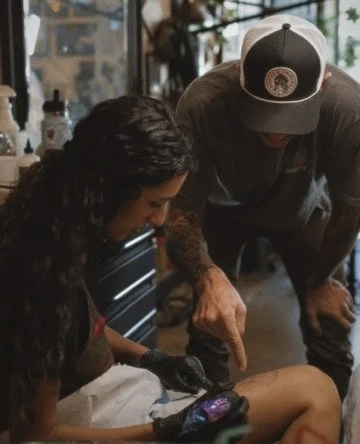 A person getting a tattoo on their arm at a tattoo shop, while a tattoo artist points at the tattoo they are working on.