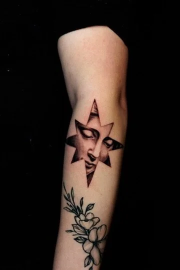 Tattoo of a woman's face within a star-shaped design on a person's arm, accompanied by a floral design below.