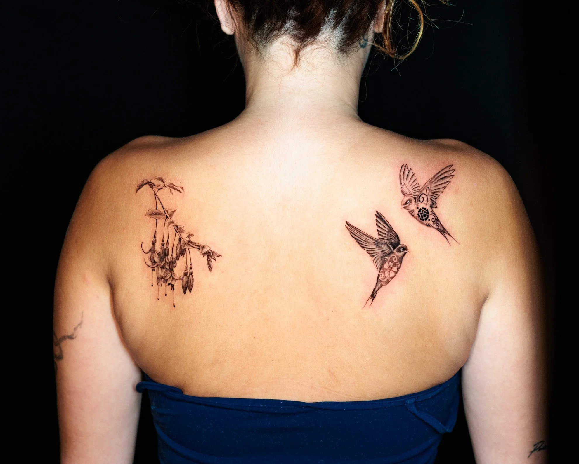 A person's back shows tattoos of hummingbirds and hanging floral designs on the shoulder.