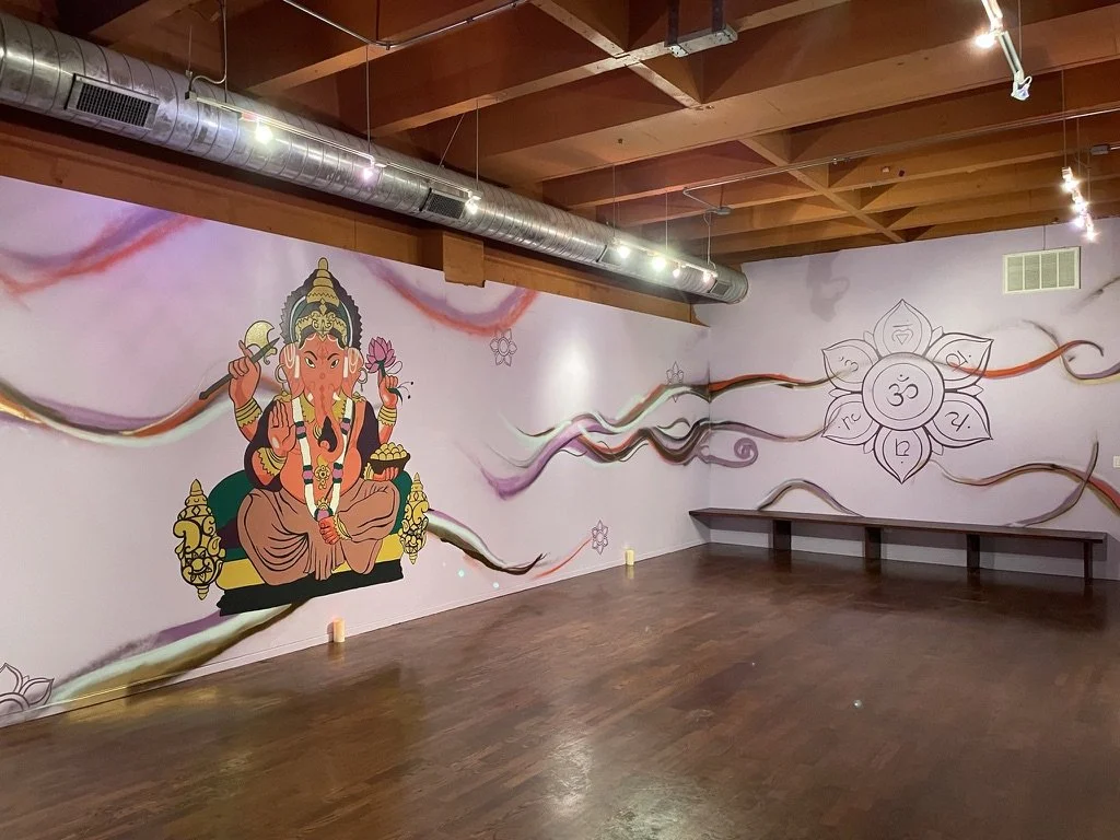 A mural featuring Lord Ganesha, an elephant-headed Hindu deity, sitting with one leg folded and holding various objects, with colorful flowing lines and a chakra with petals on the wall.