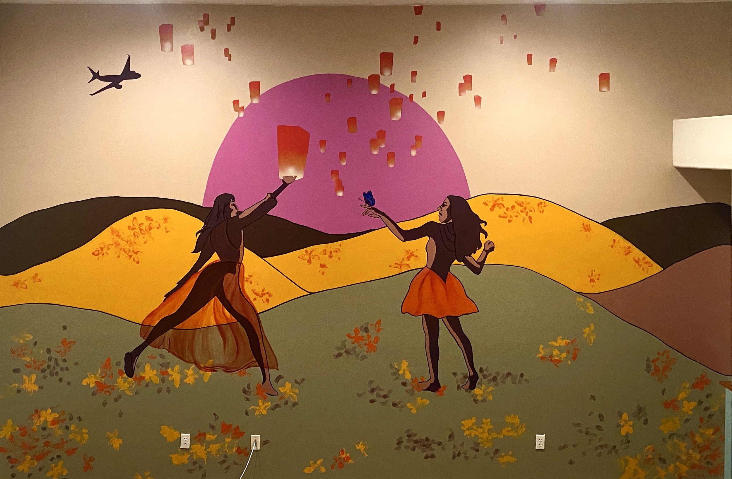 Wall mural depicting two women with long dark hair and orange skirts, with a pink sun or moon in the background, orange and yellow rolling hills, and a flying bird, with paper lanterns floating in the air.