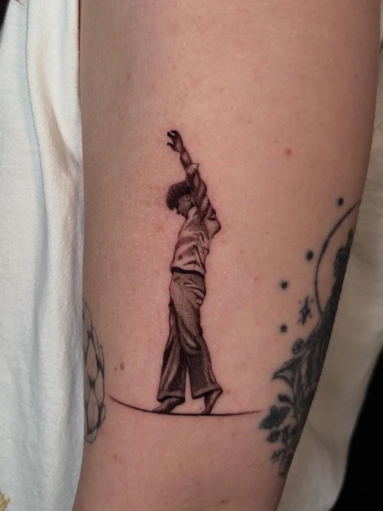 Black and gray tattoo of a person standing with arms raised above the head on a person's upper arm.