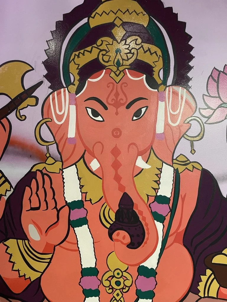 Colorful illustration of Lord Ganesha, the Hindu elephant-headed deity, shown holding a lotus flower in one hand and making a gesture with the other. He is adorned with jewelry, a crown, and traditional accessories, with a pink color skin tone.