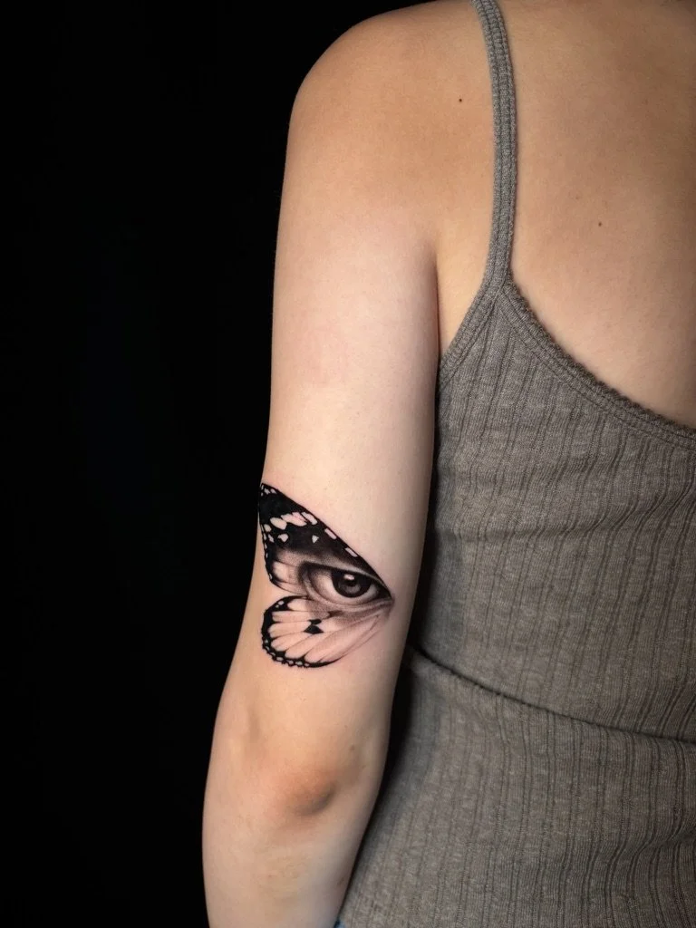 A person's upper arm tattoo of a butterfly with a realistic eye in the center of its wing.