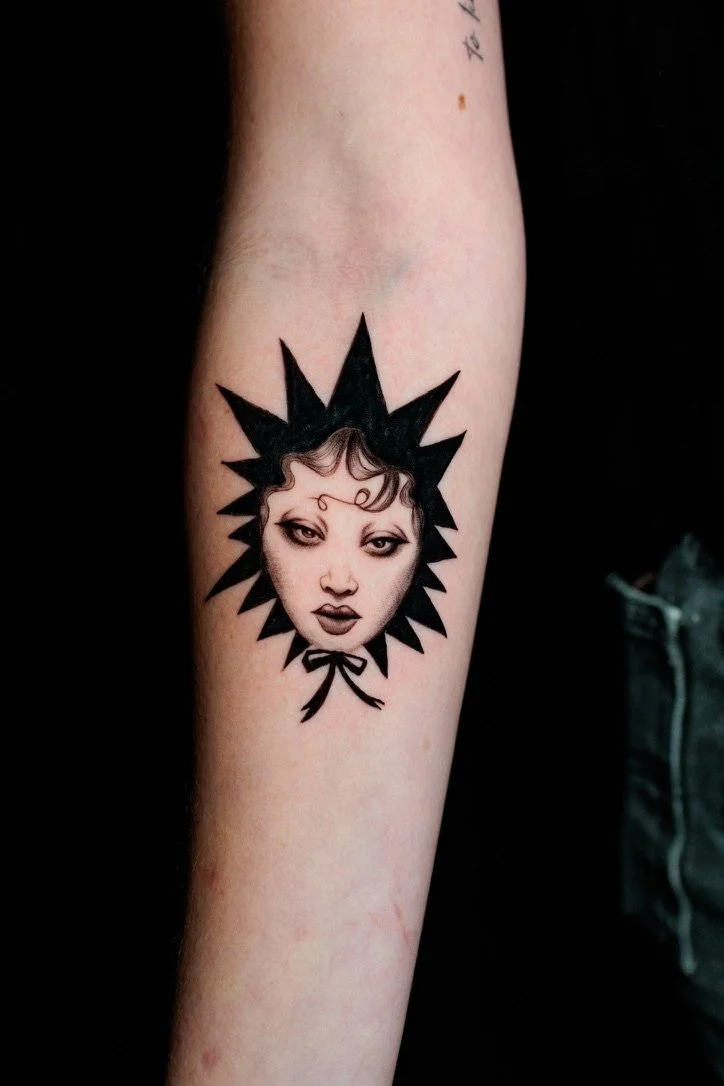 Tattoo of a woman's face with dark makeup and curled hair, framed by a black starburst shape with a bow at the bottom.
