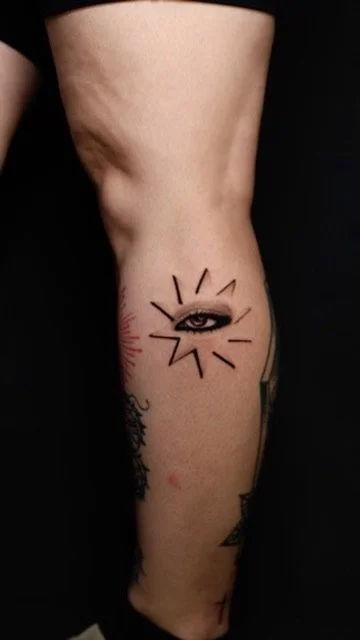 Tattoo of a stylized eye with lines radiating outward on a person's leg.