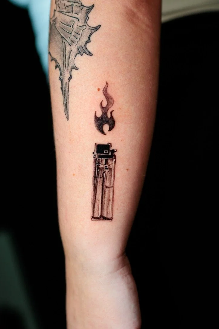 Tattoo of a lighter with a flame above it and a flame-shaped design, located on the inner forearm.