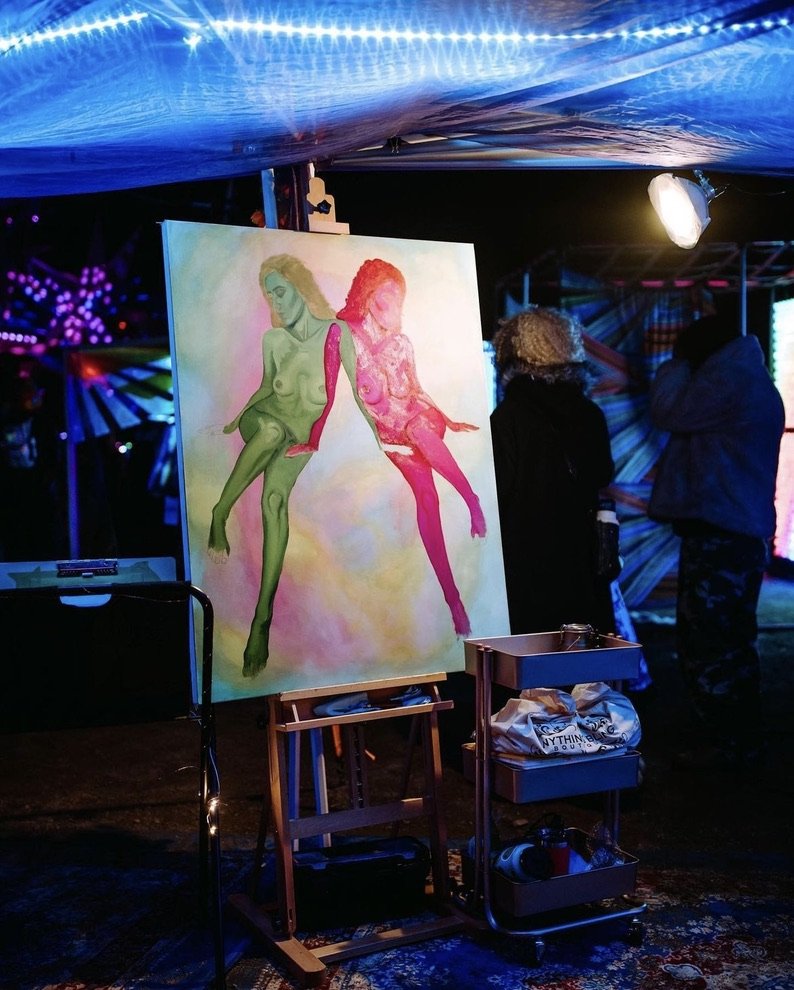 Colorful painting of two women, one green and the other pink, sitting back-to-back with long flowing hair, displayed at an art event under blue and purple lighting.