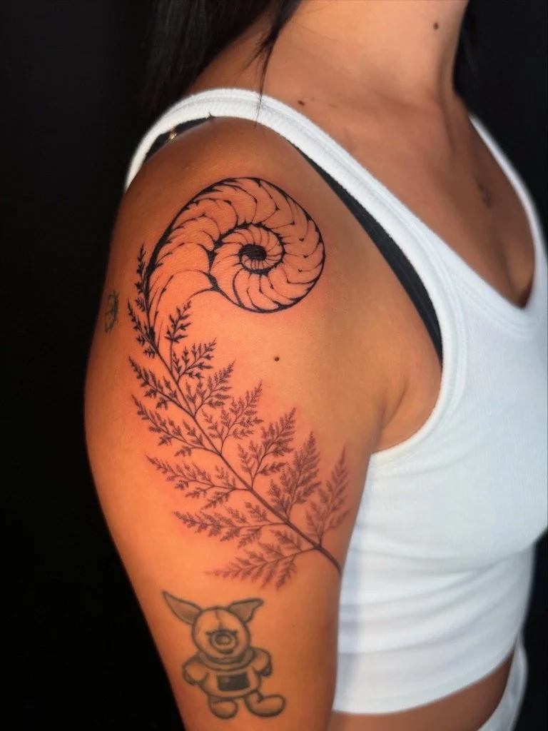 Tattoo of a spiral shell and fern on upper arm, with a small bear-shaped character below.