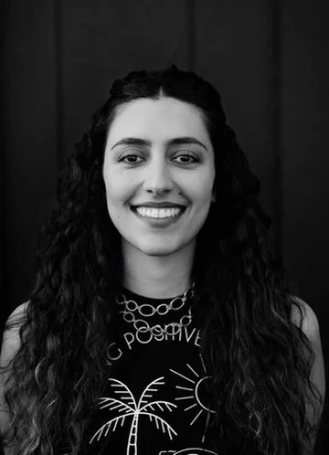 A smiling young woman with curly hair, wearing a sleeveless top with a sun and palm tree graphic, and layered necklaces, standing in front of a dark background.