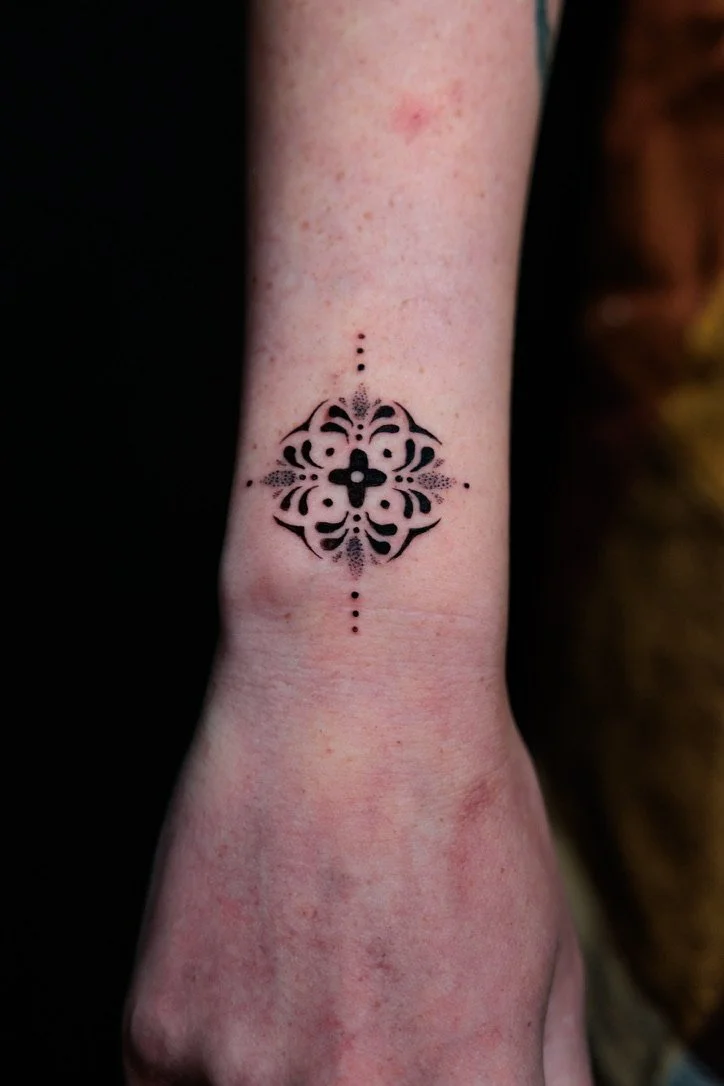 Tattoo of an ornate black symmetrical design with dots above and below on a person's wrist.