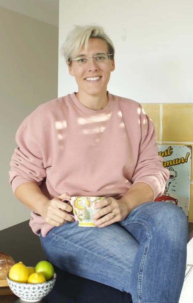 Christine is wearing a pink jumper and jeans, sitting on a bench in the kitchen with a cup of coffee.