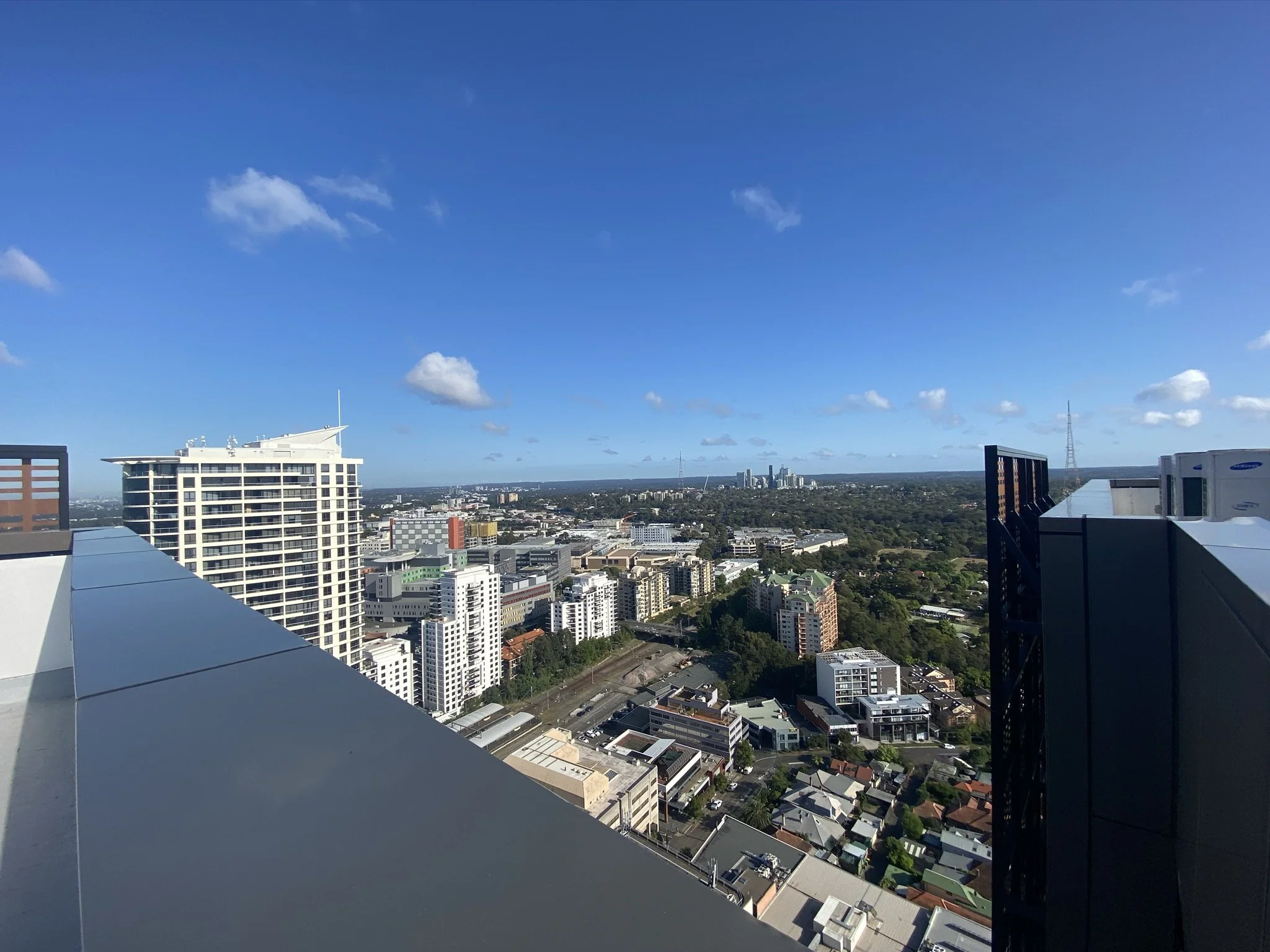 View of the north shore from a high-rise building in st leonards with a clear blue sky, a few clouds, and various buildings, including a distinctive white building with an observation deck, in the distance.