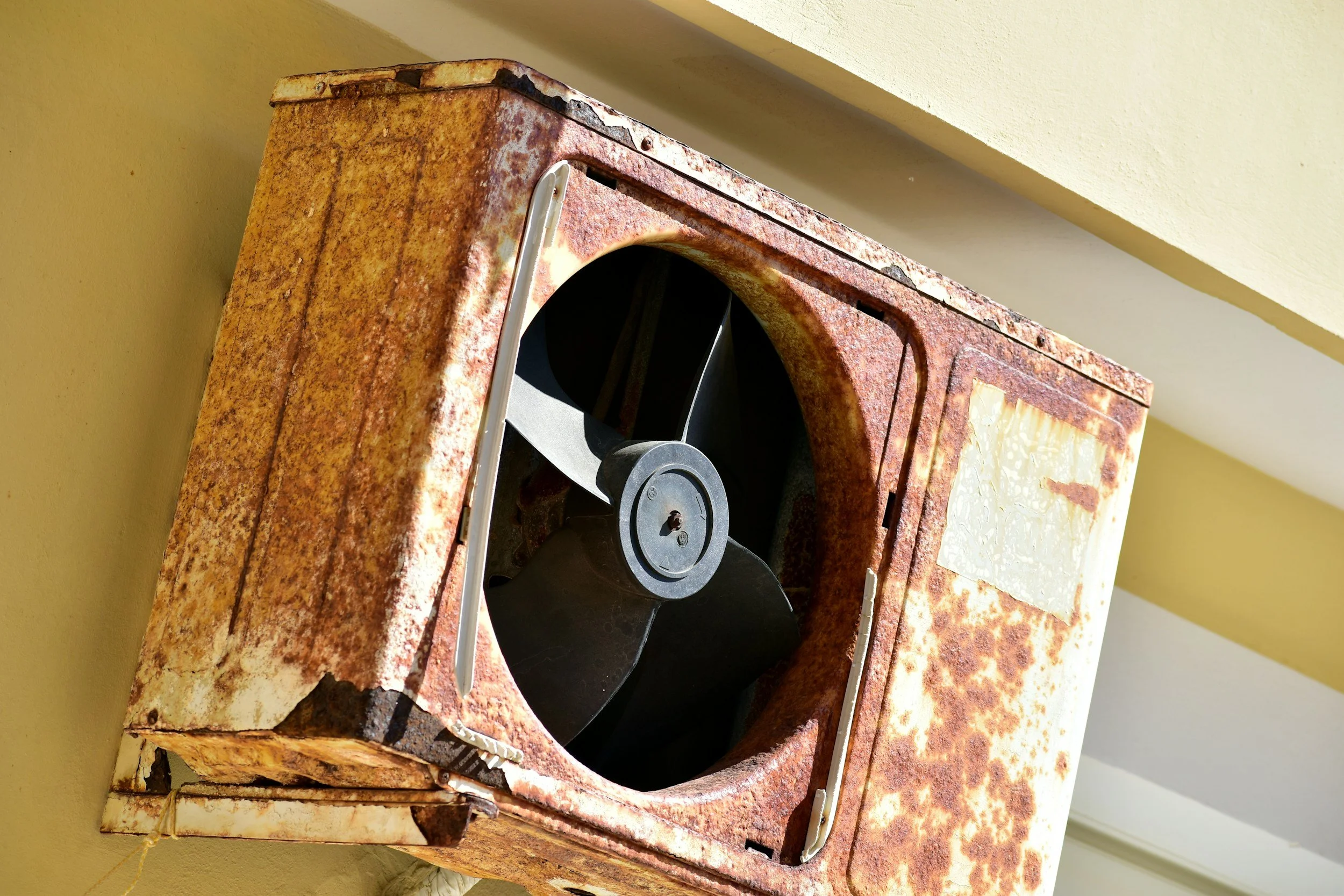 A rusty, old condensing unit, with a black fan inside, mounted on a yellow wall.