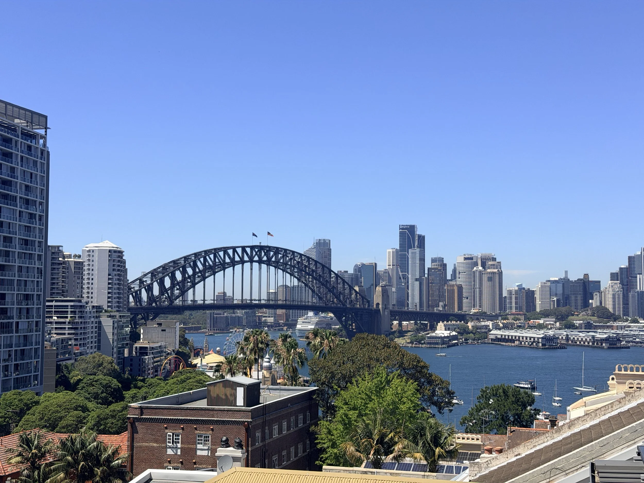A cityscape featuring the Sydney Harbour Bridge, high-rise buildings in the cities CBD, Sydney harbour, and trees on a sunny day.