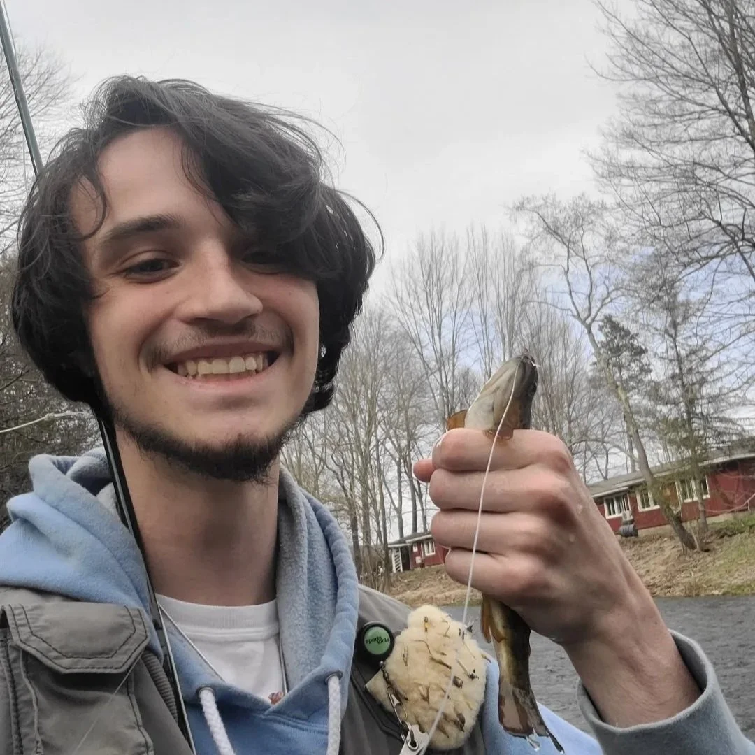 A young man with dark wavy hair, smiling and holding a fish he caught. He is outdoors near a river with leafless trees and houses in the background.