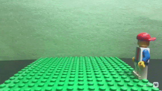 A LEGO figure dressed in a blue shirt, gray pants, and a red cap stands on a black surface next to a large green LEGO baseplate against a green background.