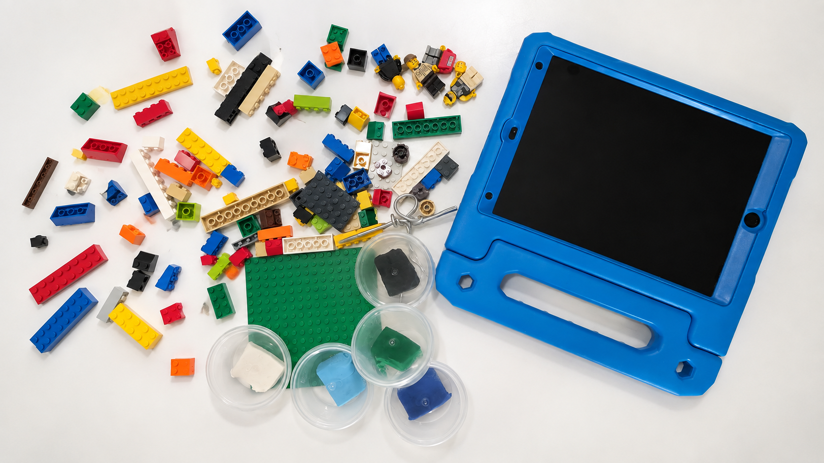 A blue kids' tablet case with a black tablet inside, scattered LEGO pieces in various colors and sizes, small containers with colored clay or putty, and a green LEGO baseplate on a white surface.