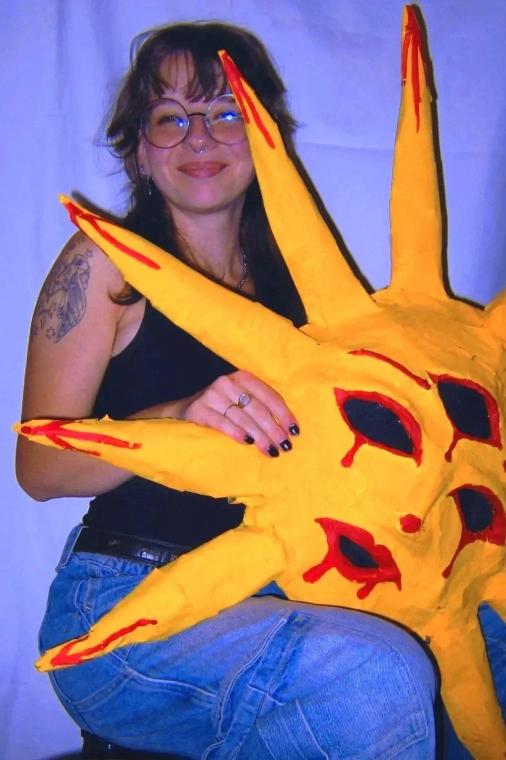 A long-haired person with glasses and tattoos is smiling and holding a large, bright yellow, sun mask with red and black face details on it.