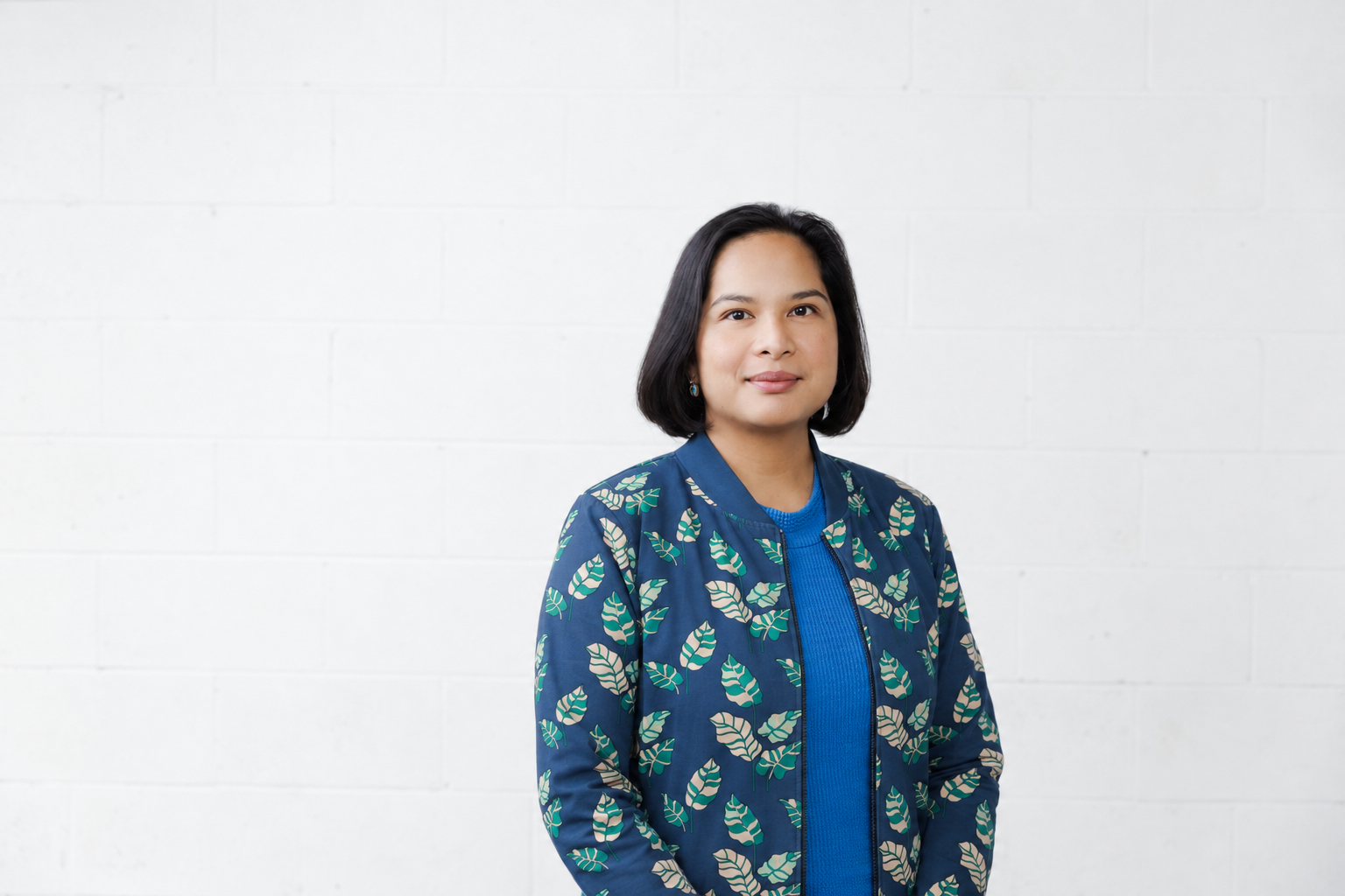 A woman with shoulder-length black hair wearing a blue jacket with leafy patterns and a blue top, standing against a white wall.