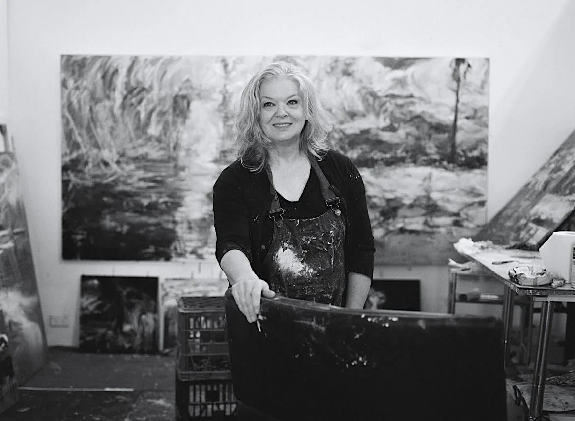 Laura Matthews in her studio standing in front of her large landscape painting, wearing an apron covered in paint and holding a palette.