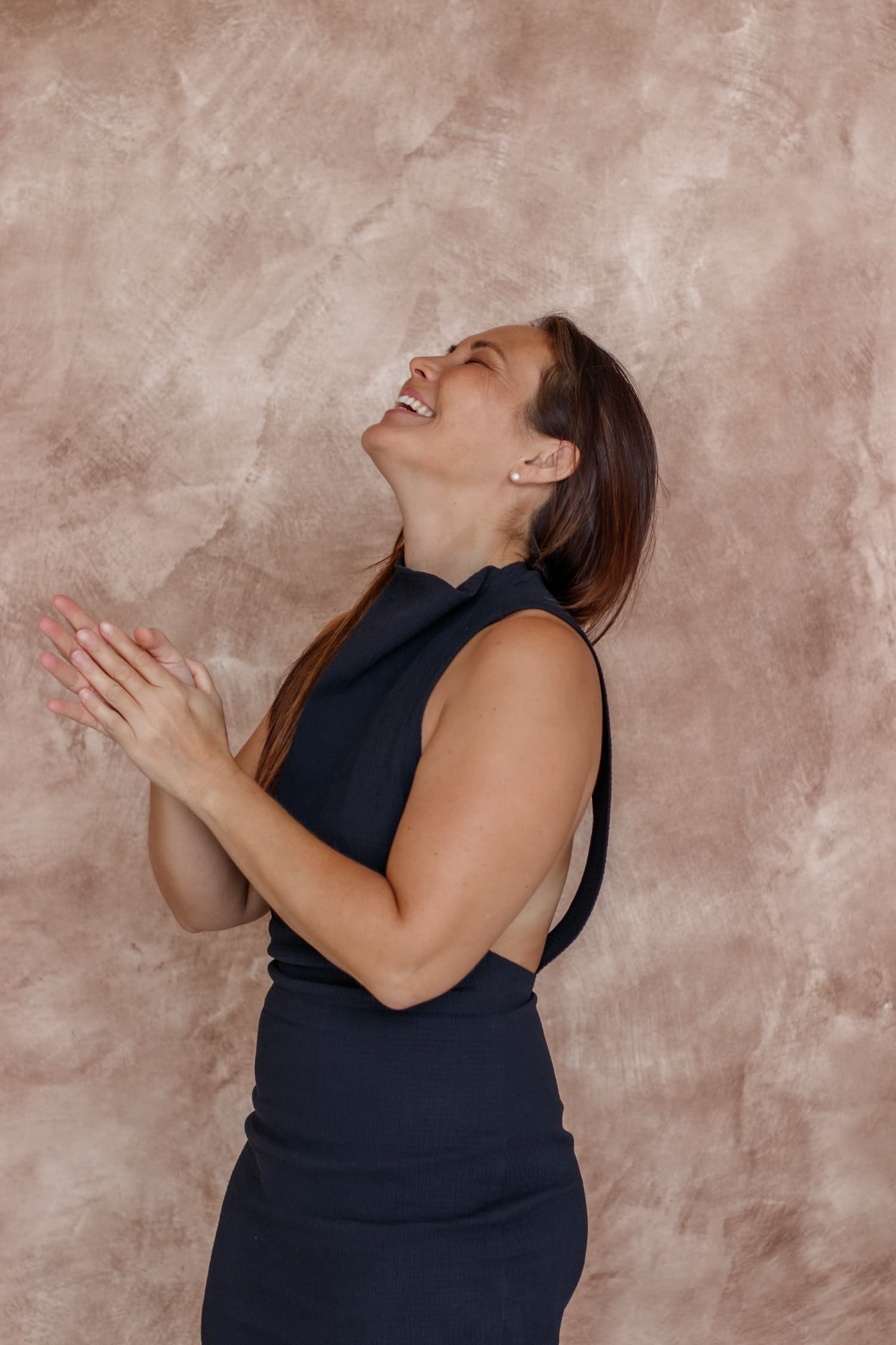 Side view of a woman smiling with eyes closed, clasping her hands together, wearing a black sleeveless dress, standing against a textured beige background.