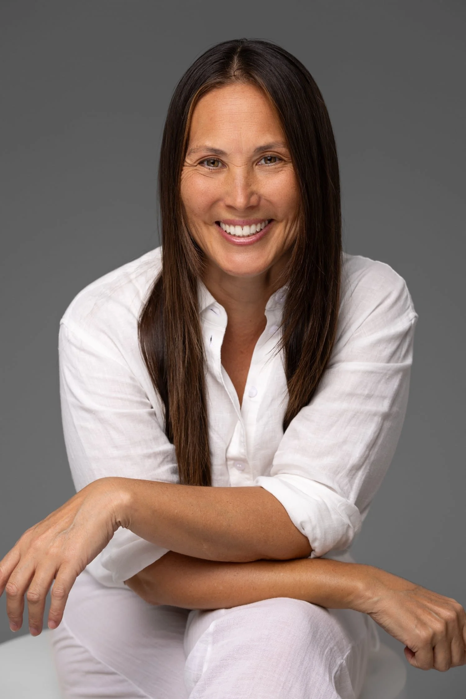 A woman with long dark hair wearing a white shirt, smiling, sitting with her arms crossed against a gray background.