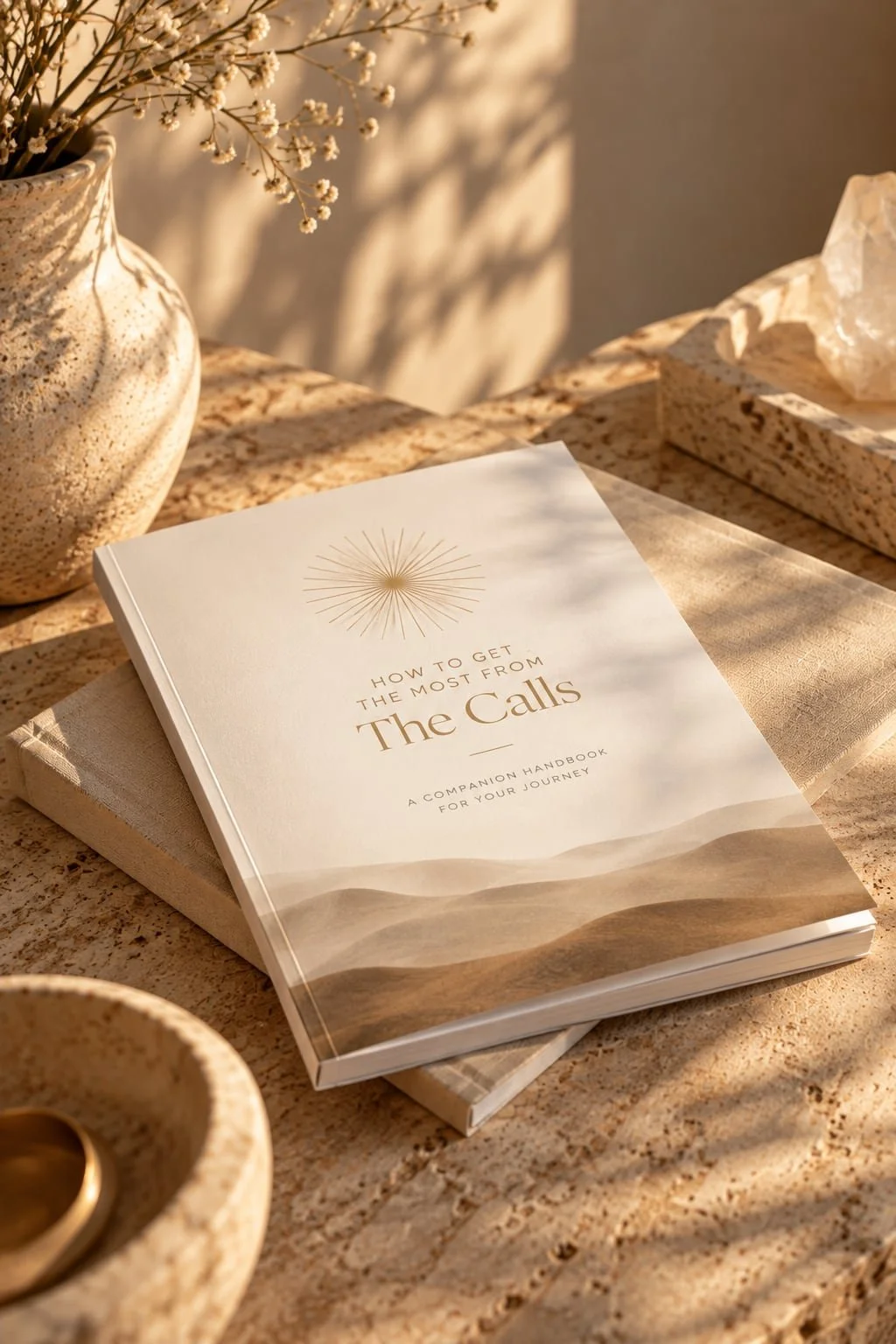 A book titled 'How to get the most from The Calls' resting on a second book on a stone tabletop. There is a vase with white flowers, a small bowl with a gold object, and a tissue box nearby, with warm sunlight casting shadows.