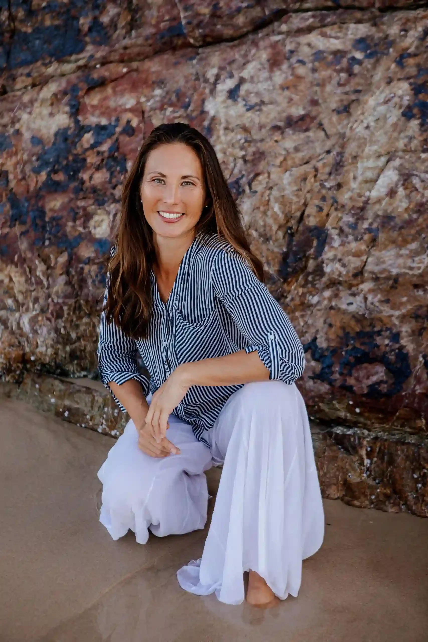 A woman with long brown hair and a striped shirt with rolled up sleeves sitting on the sand at the beach, smiling at the camera with a rocky wall in the background.