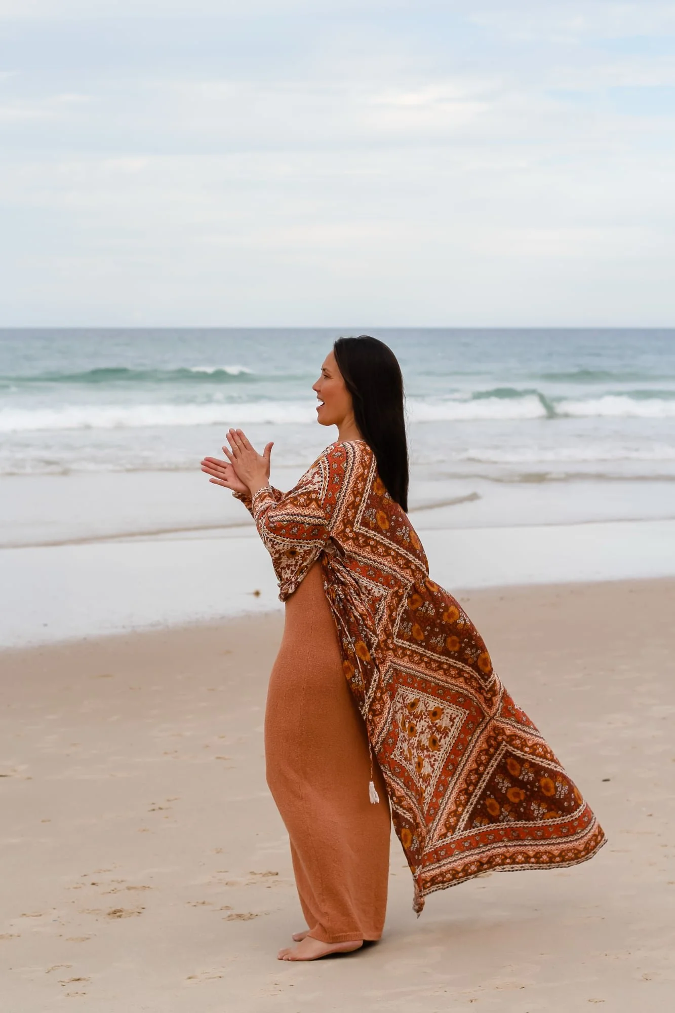 A woman standing barefoot on a beach, facing the ocean, wearing a patterned shawl and light brown pants.