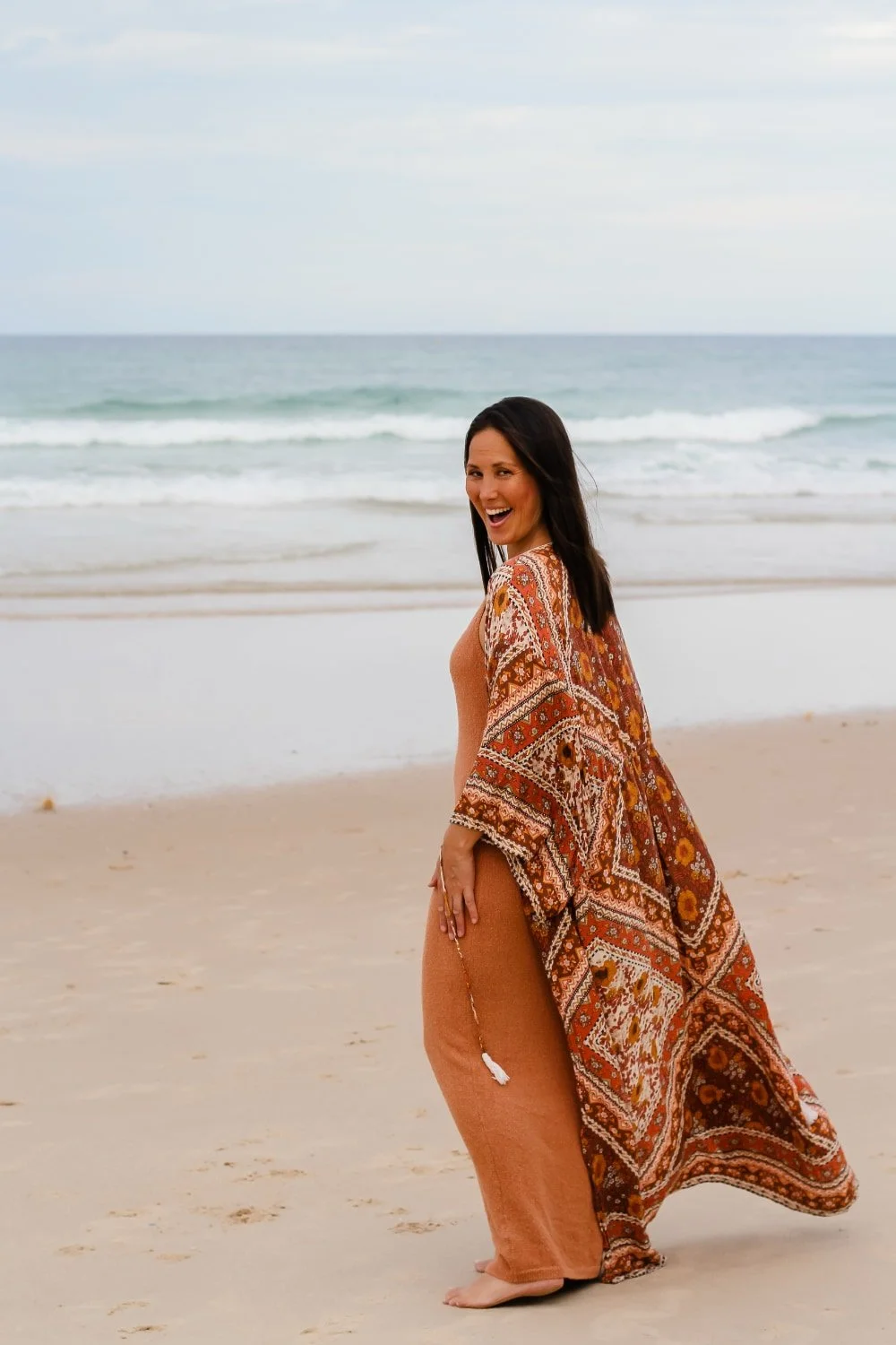 A woman in a rust-colored outfit and patterned cover-up standing on a sandy beach with ocean waves in the background, smiling and looking at the camera.