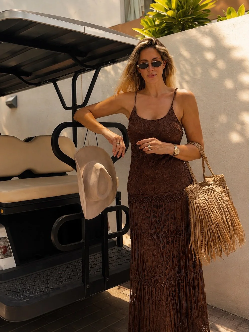 Woman in a brown lace dress holding a straw bag, sunglasses, and a watch, standing next to a golf cart with hats hanging on it, outdoors with sunlight and greenery in the background.