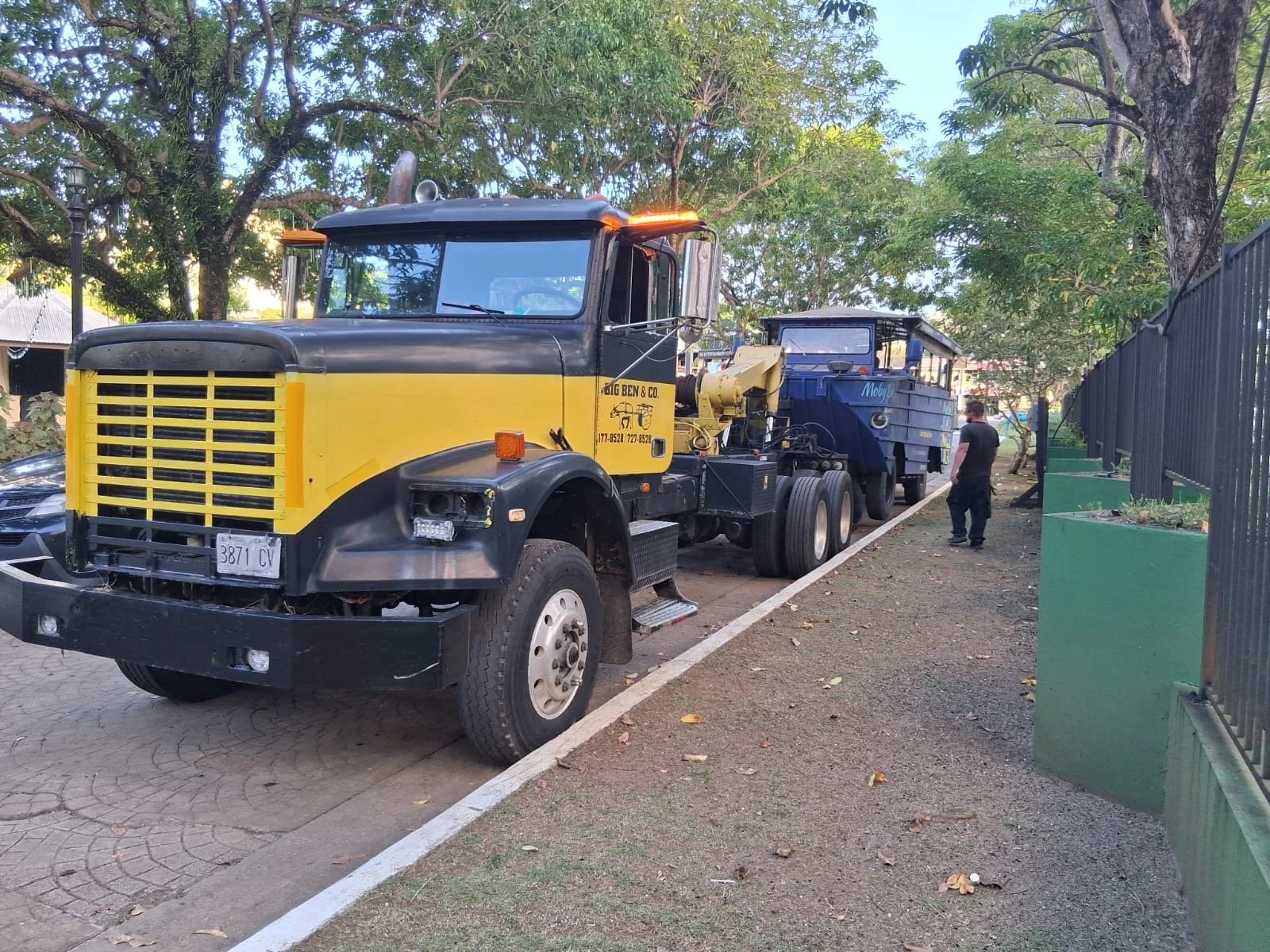 A large black and yellow truck parked on the side of a street, with a person standing nearby. Trees and a fence are in the background.