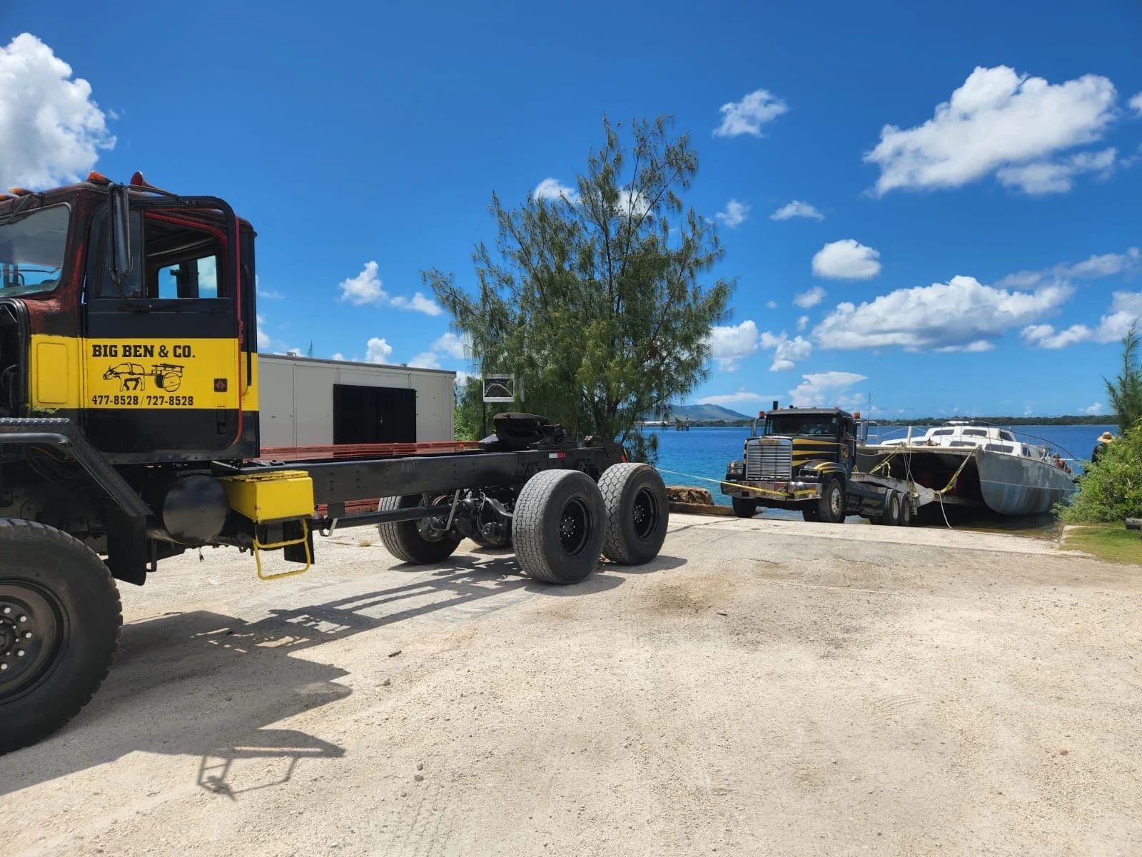 A tow truck pulling a large boat out of the water at a dock, with a tree and blue sky with clouds in the background.