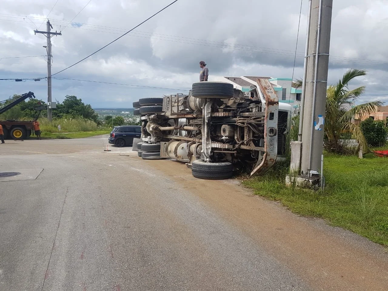 An overturned semi-truck lying on its side at the intersection, with a man standing on top of the truck, and a few people near the scene. There are also some vehicles parked nearby and utility poles with wires on the side of the road.