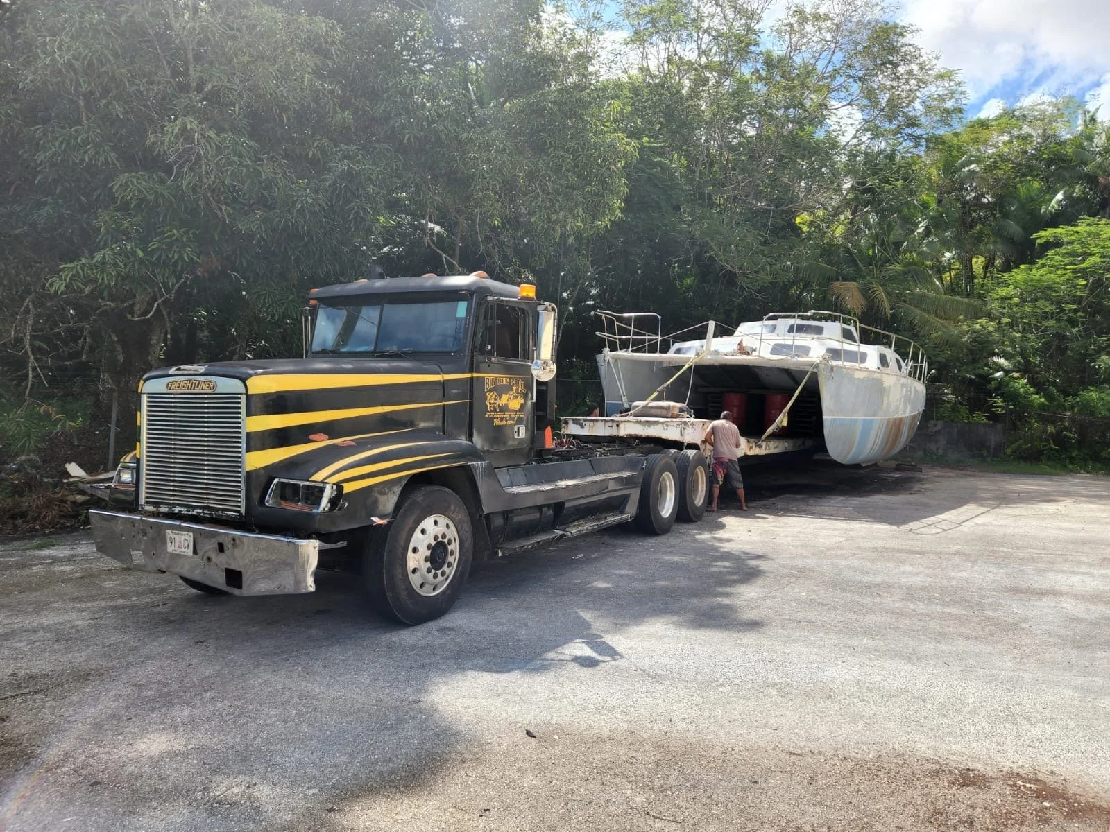 A black semi-truck pulling a white and blue yacht on a trailer, parked on the side of a road with green trees behind.