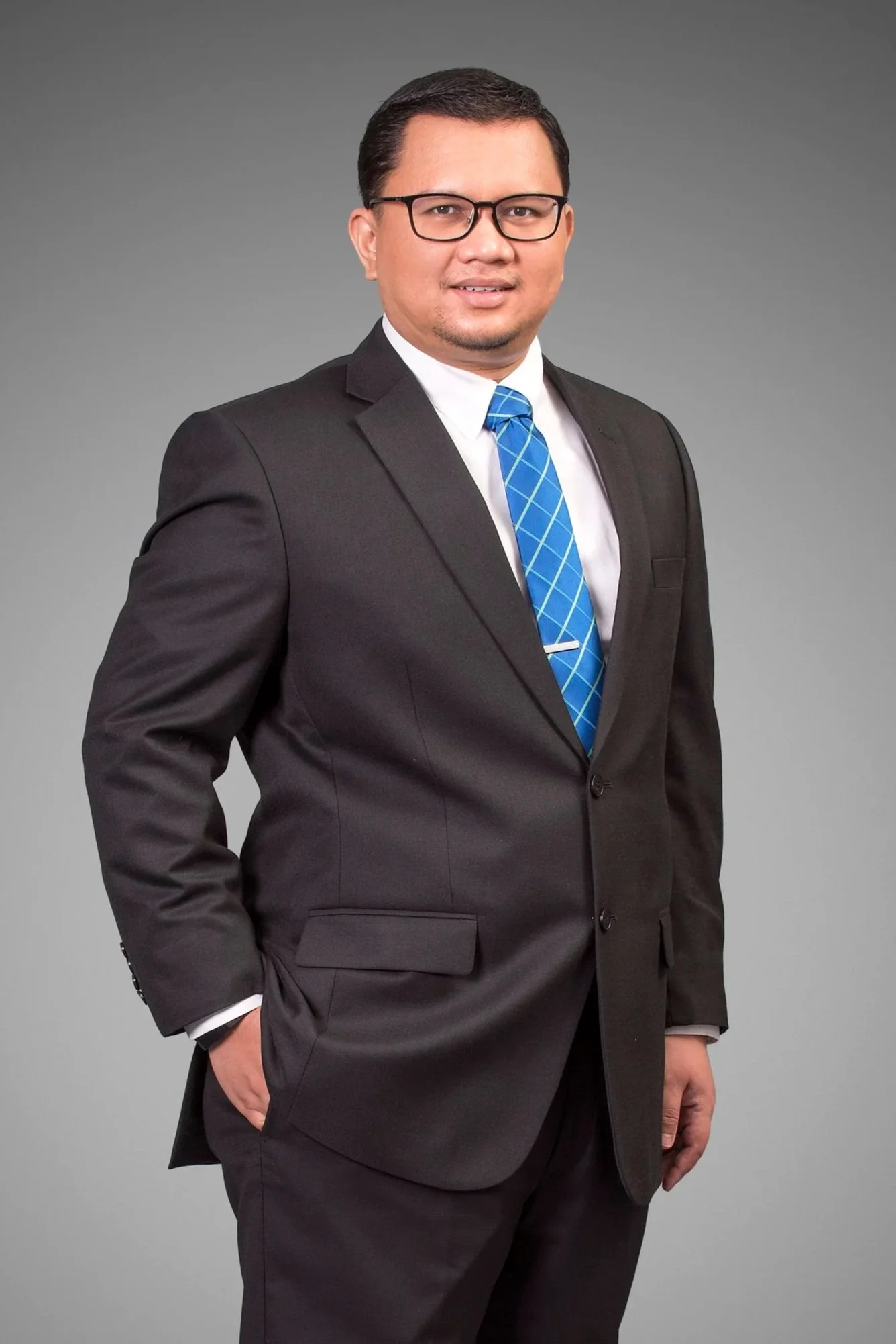 A man in a dark suit, white shirt, blue plaid tie, and glasses poses against a gray background.
