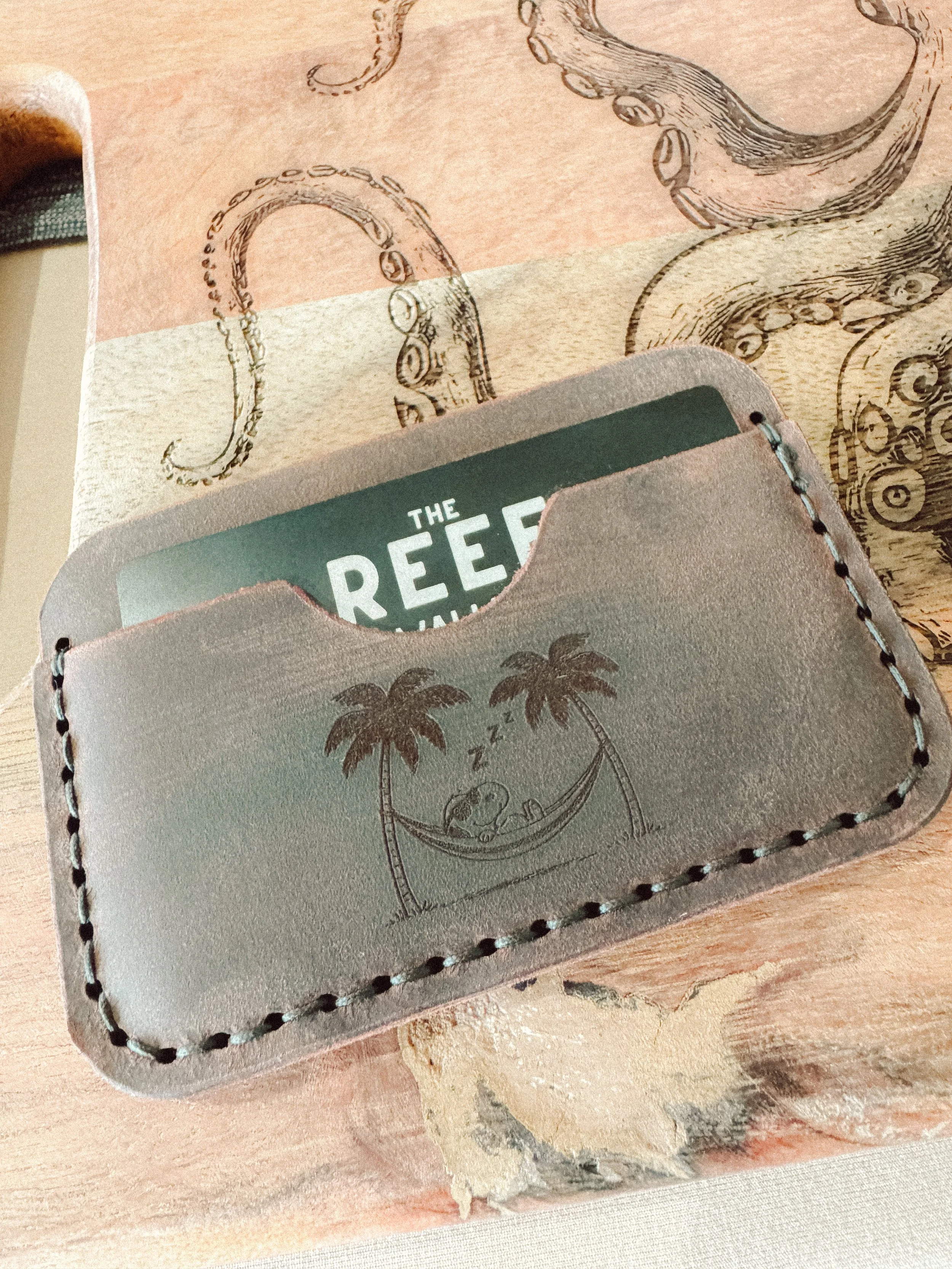 Close-up view of a gray leather pocket with a black and white illustration of a snow scene, palm trees, and sleeping Snoopy, with a partial glimpse of a box labeled 'THE REEF' in the pocket.