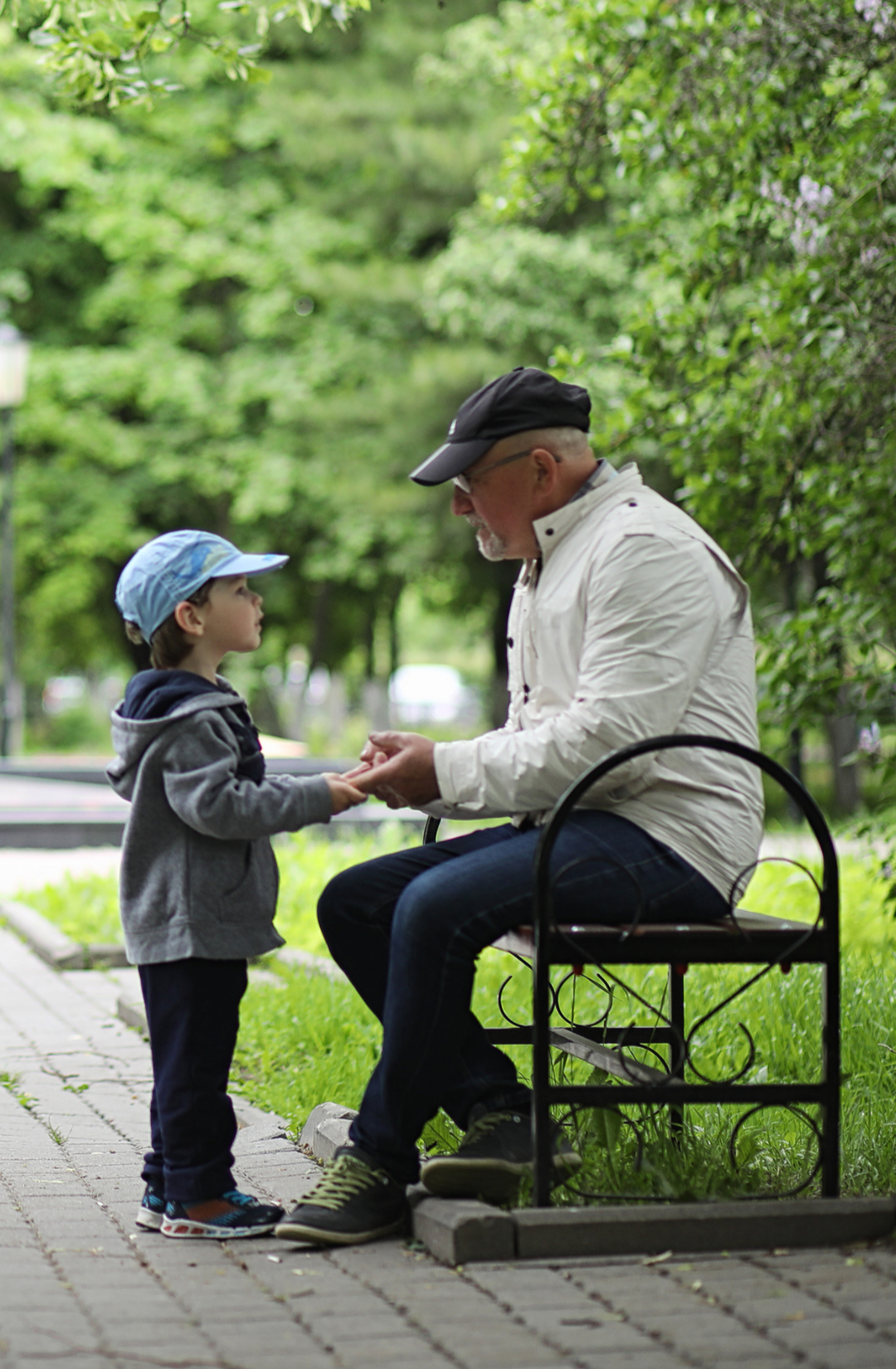 A man sitting on a park bench holding hands with a young boy, engaging in a conversation amidst green trees and a paved walkway.