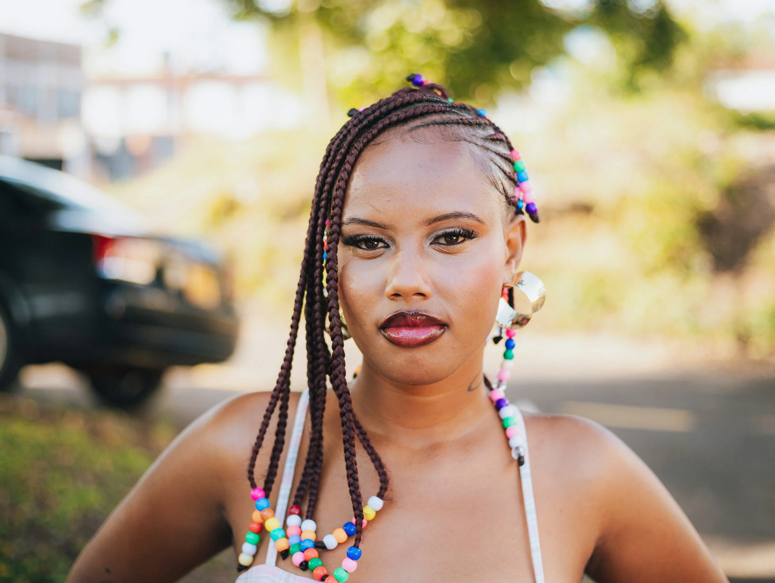 A young woman with braided hair and colorful beads wearing large earrings, outdoors on a sunny day.