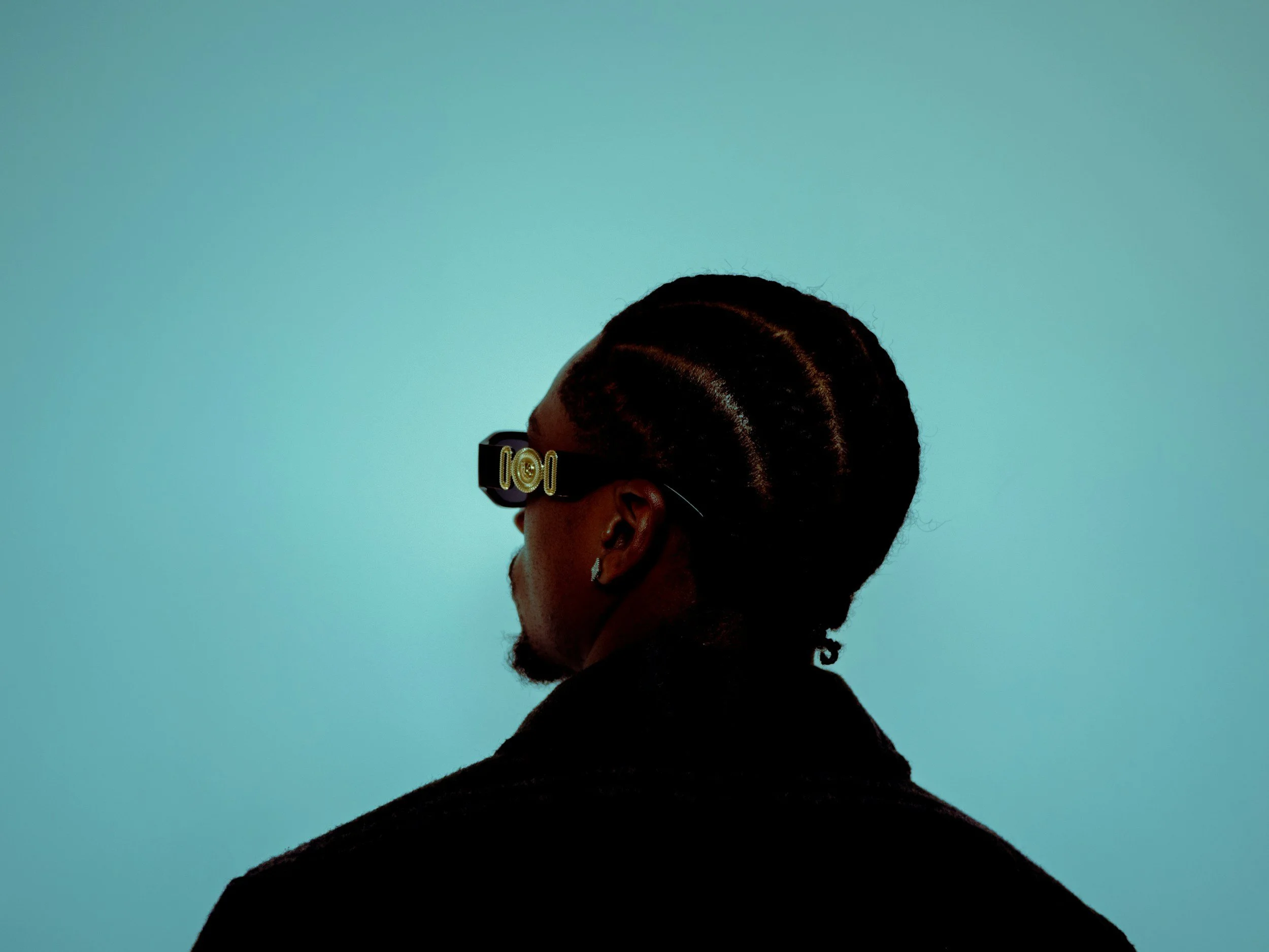 Profile of a woman with braided hair, wearing sunglasses with a decorative gold logo, against a solid light teal background.