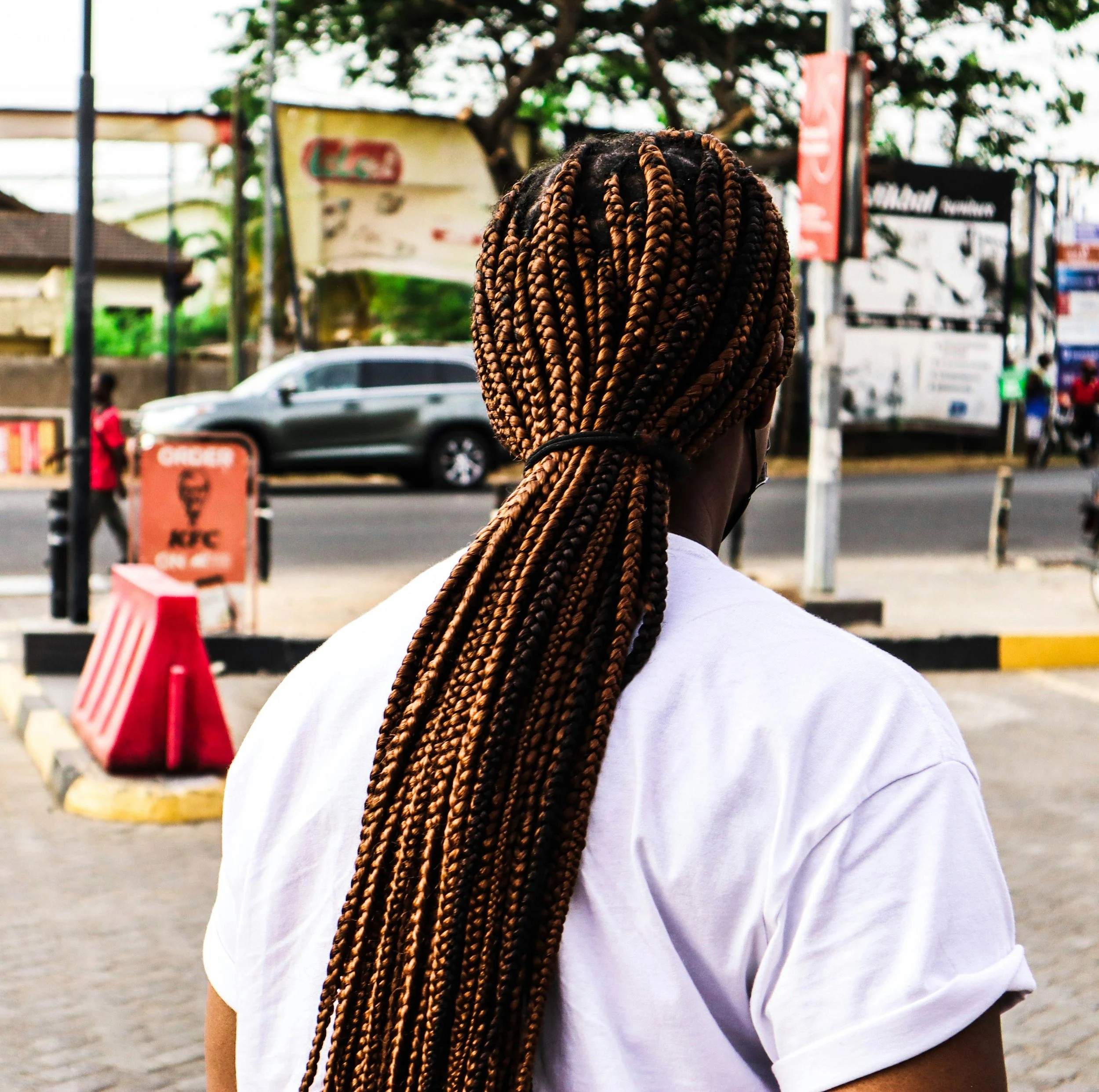 Head and shoulders of a woman with long, thick, blonde box braid hair standing outdoors in an urban setting near a street with cars and sidewalk signs.